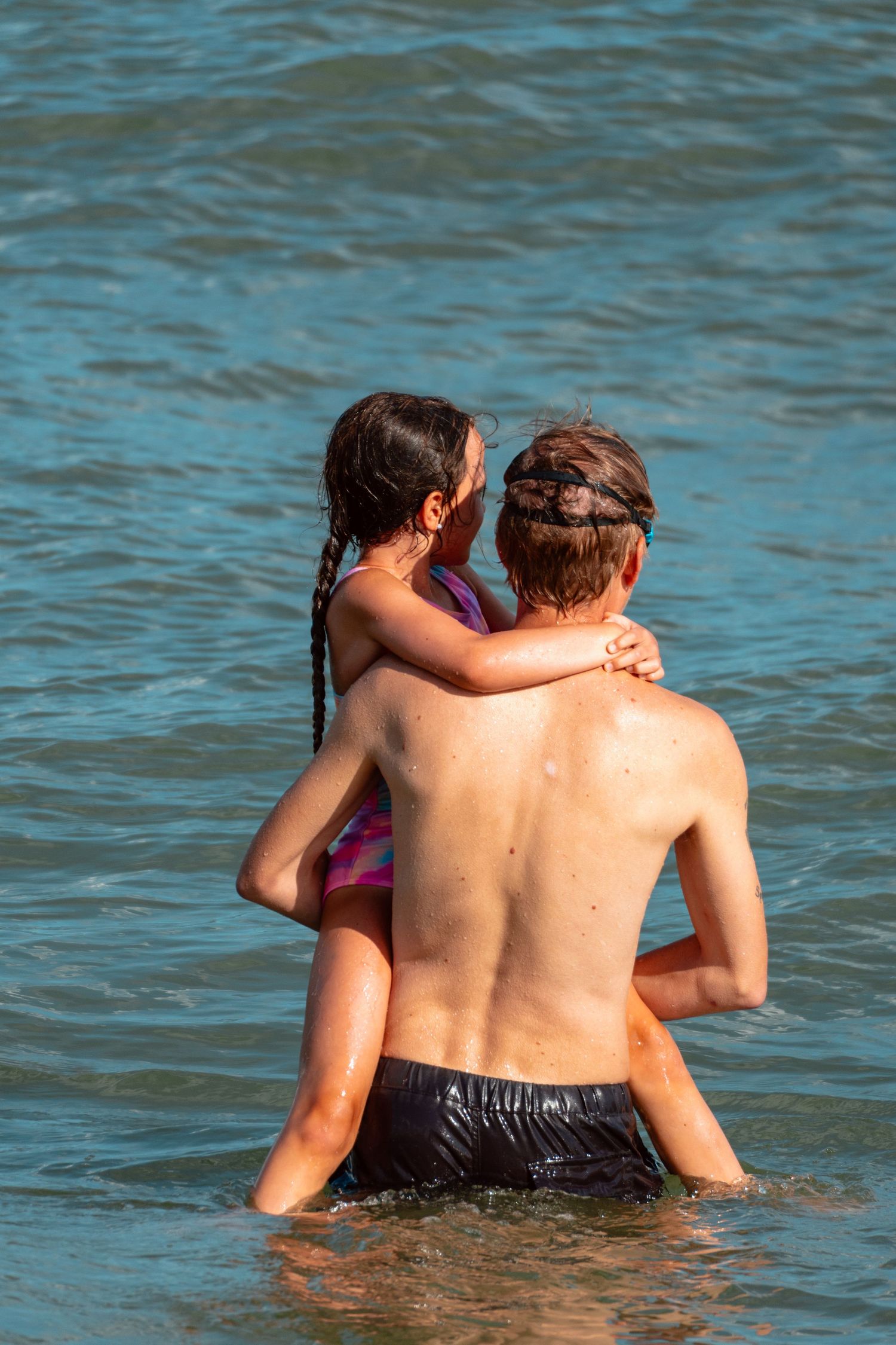 Adult carrying a young girl in a pink swimsuit on their back while wading in blue ocean water.