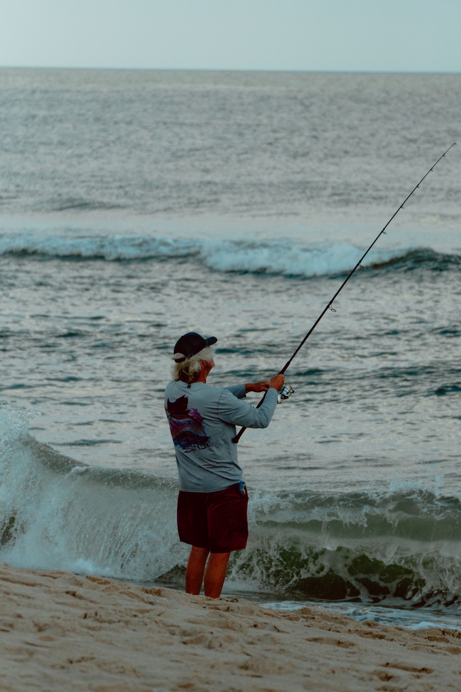Surf fisherman casting a rod into ocean waves from a sandy beach at dusk.