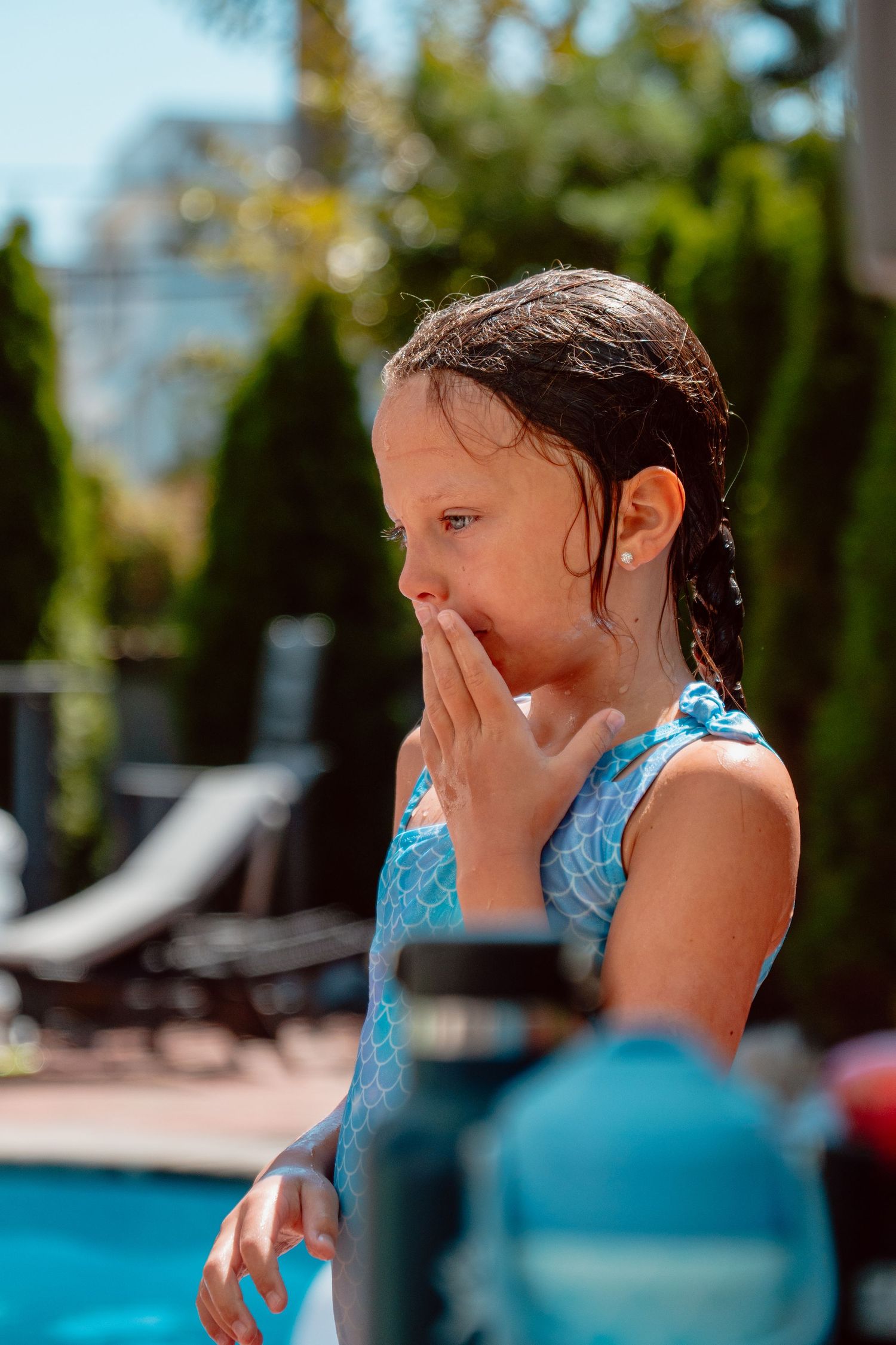 Wet-haired girl in blue swimsuit covering her mouth by a sunny backyard pool.