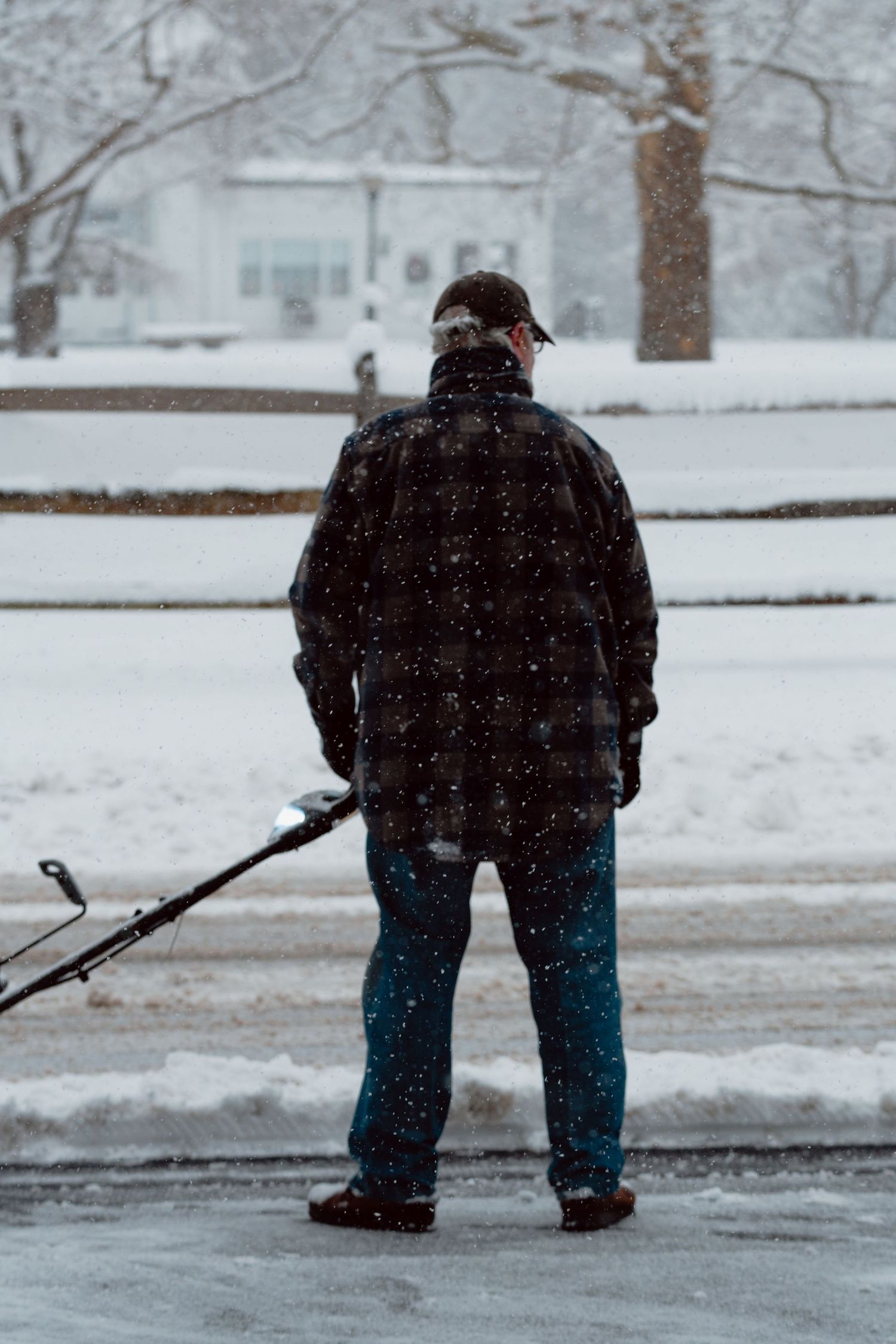 A man in a plaid jacket standing in a snowy driveway holding a snowblower during a blizzard.