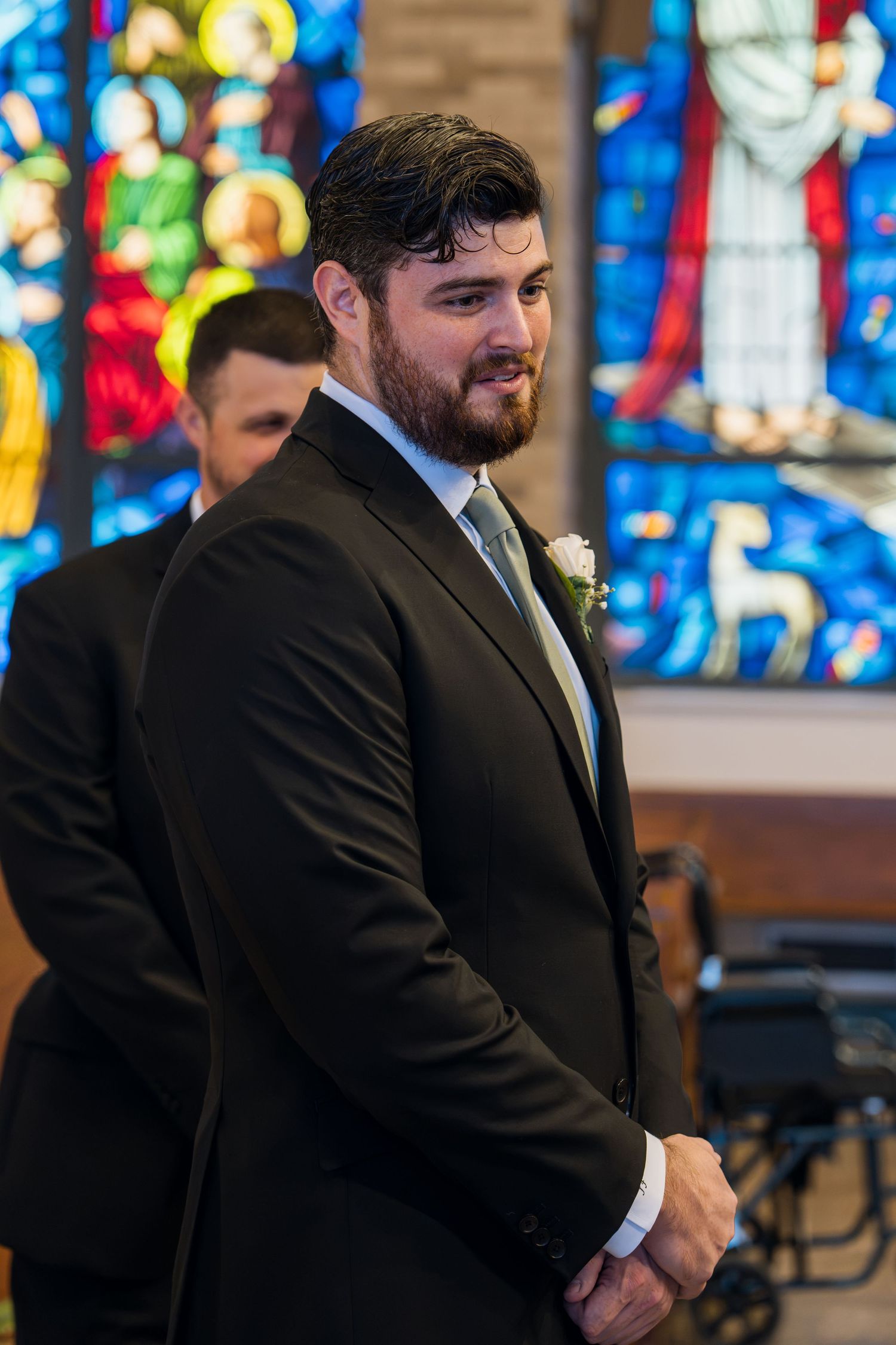 Groom in black suit with white boutonniere smiling at church altar with colorful stained glass windows behind him.