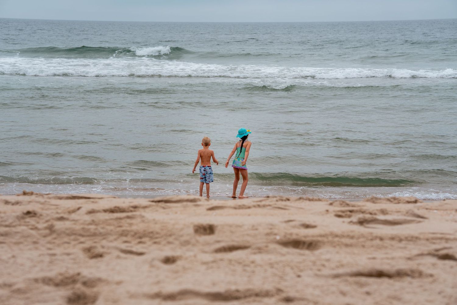 Two children holding hands at the ocean shore, standing on sandy beach with gentle waves in the background.