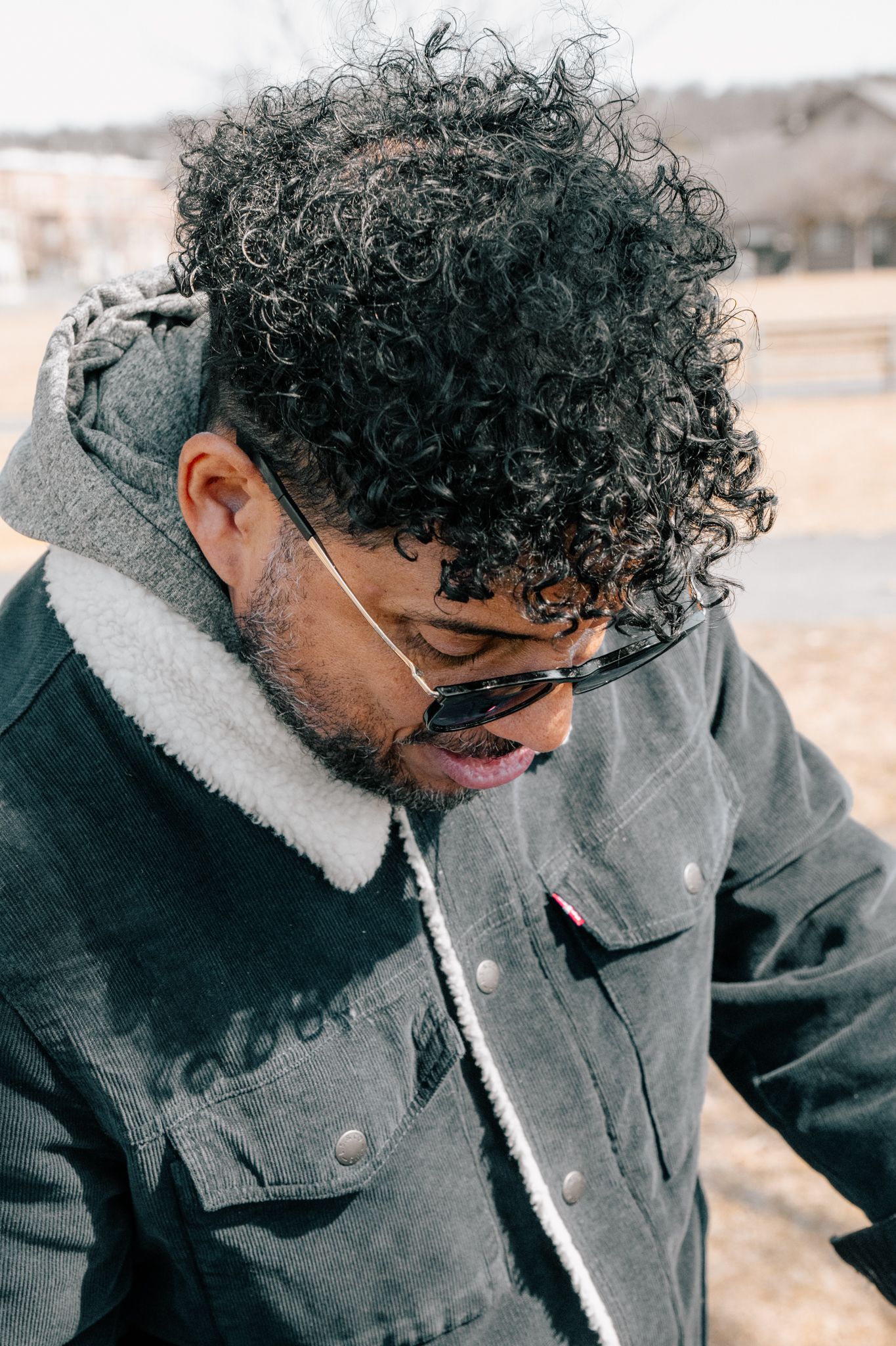 Close-up portrait of curly-haired man with sunglasses looking down in outdoor setting.