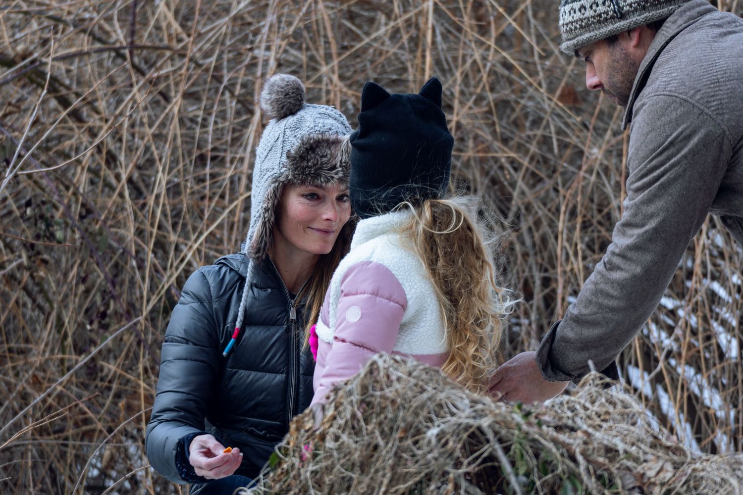 Two women and a man interacting near dry winter brush in snowy conditions.
