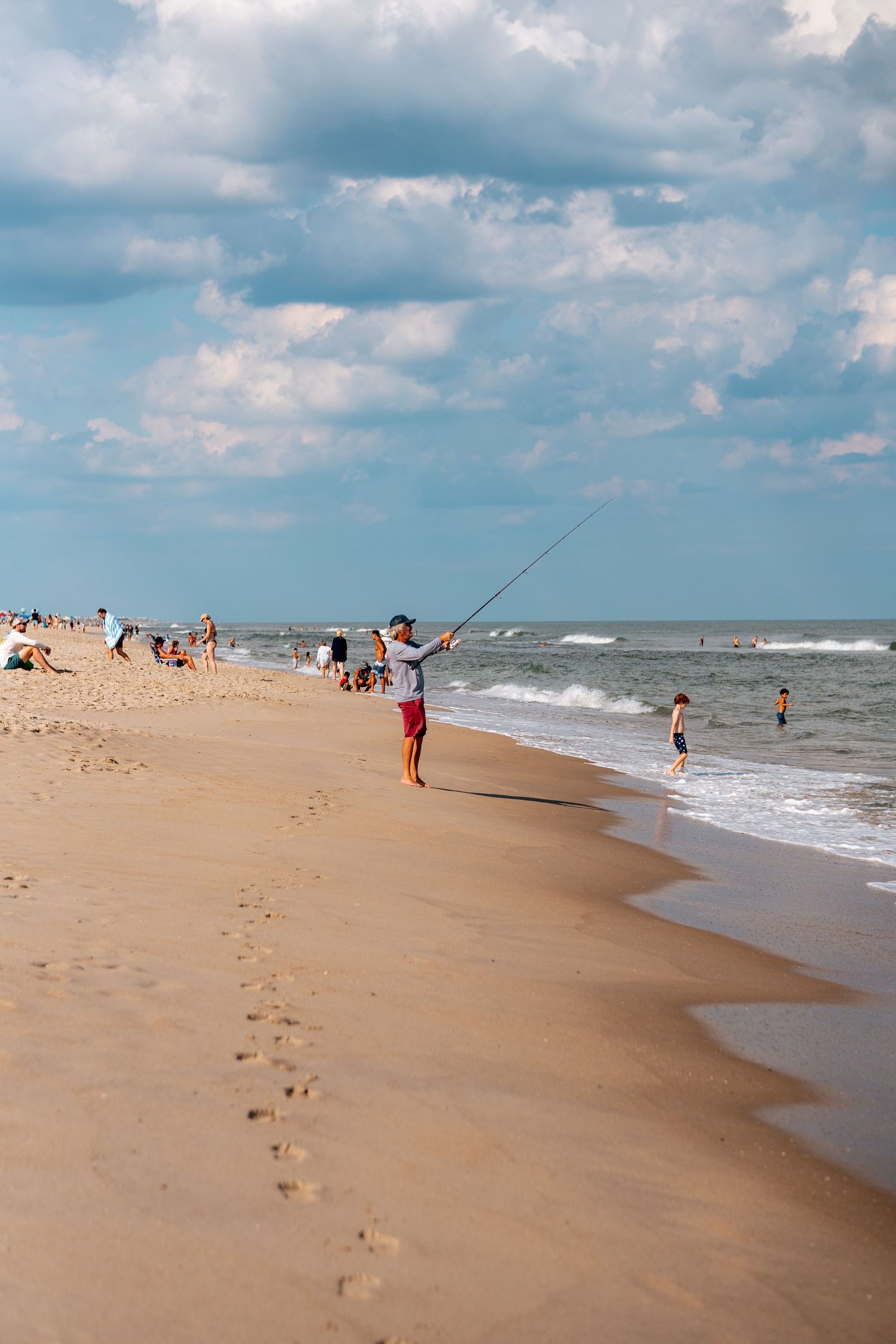 Surf fisherman casting a line on a wide sandy beach with swimmers and beachgoers nearby.