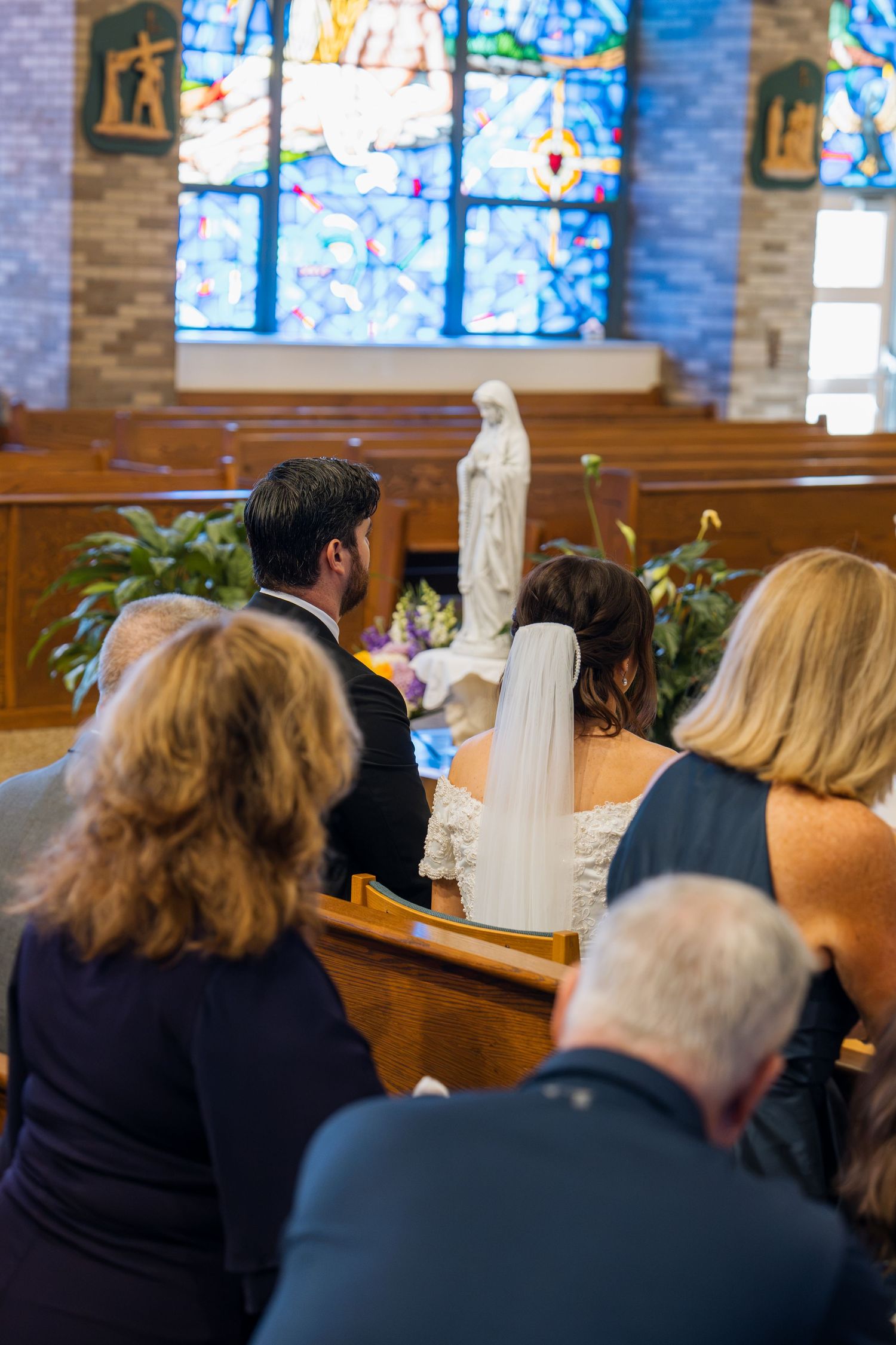 Catholic wedding ceremony with bride and groom kneeling before Virgin Mary statue, stained glass windows in background.