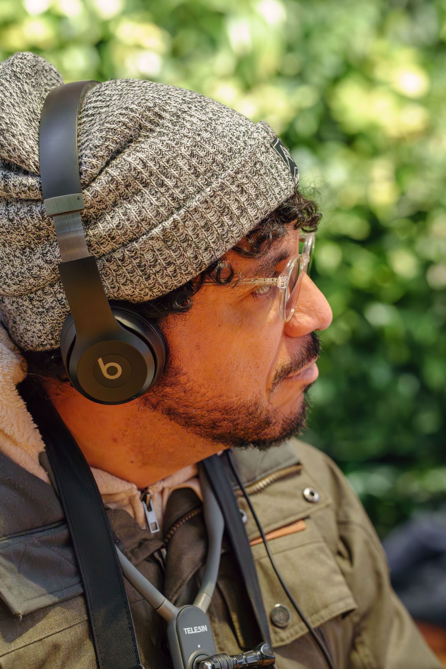 Side profile of a bearded man wearing Beats headphones and gray knit beanie against green foliage.