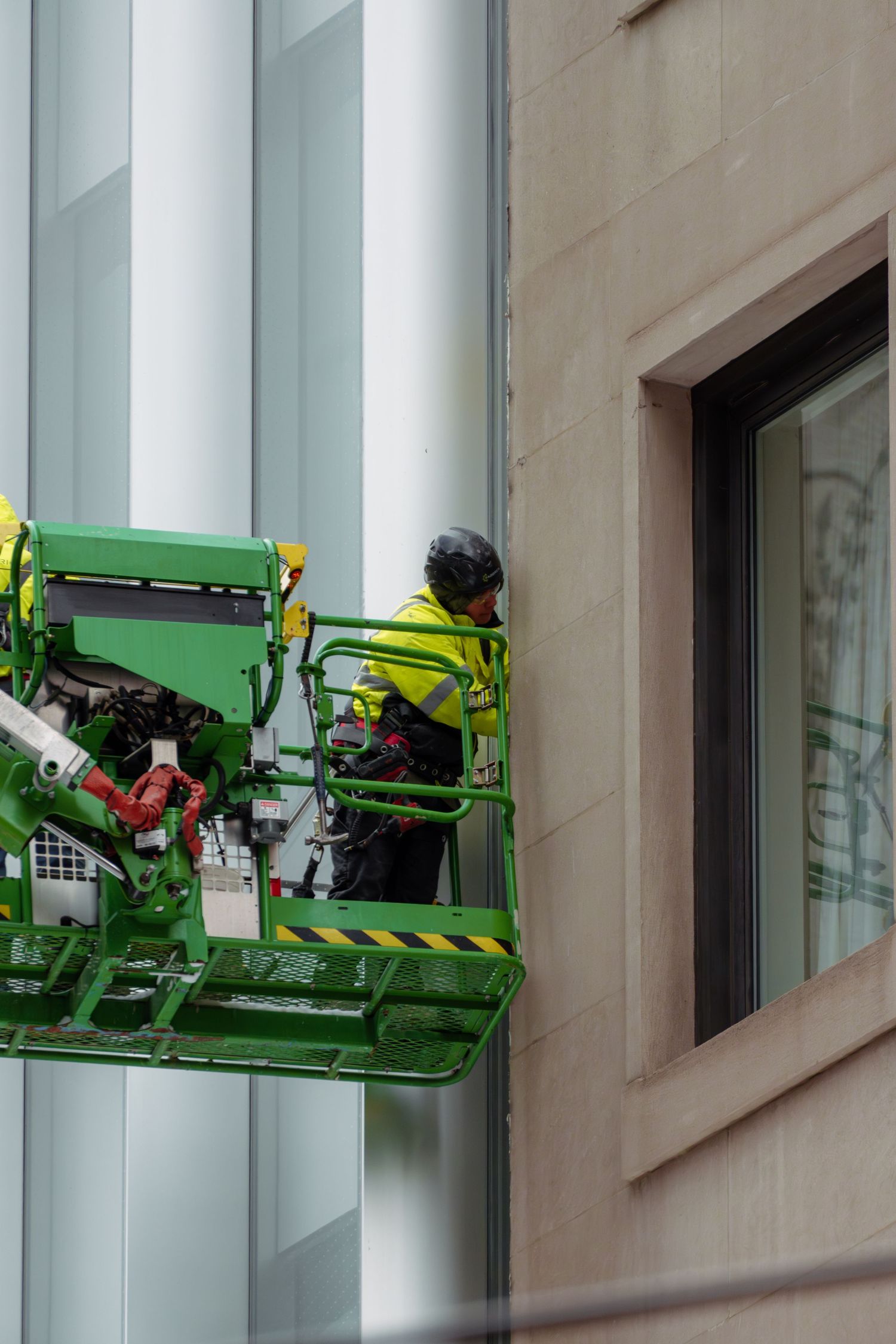 Worker in yellow safety vest on green cherry picker lift maintaining building exterior.