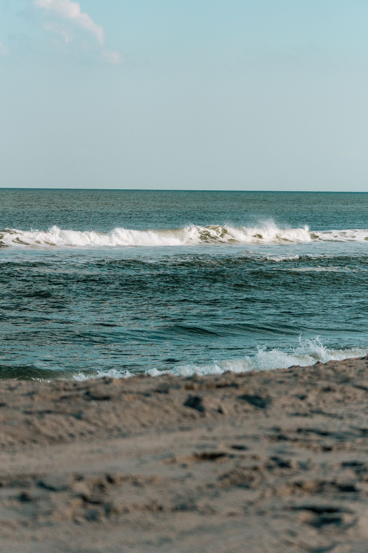 Powerful ocean waves crash onto a sandy beach shoreline with white sea foam under a light blue sky.