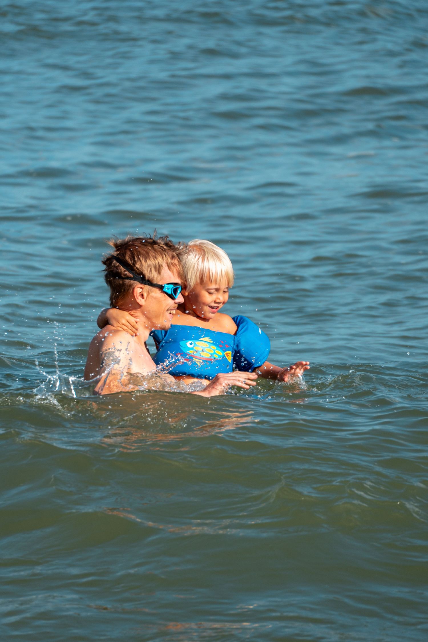 Two children playing in ocean water, with one wearing blue floaties and goggles while being hugged.