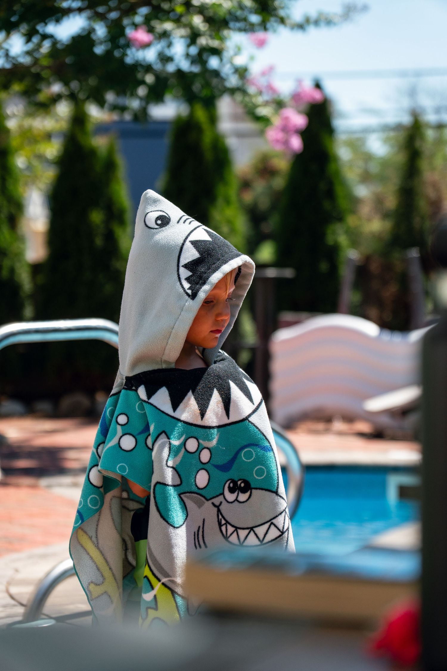 Young boy wrapped in a shark hooded towel standing beside a backyard swimming pool.