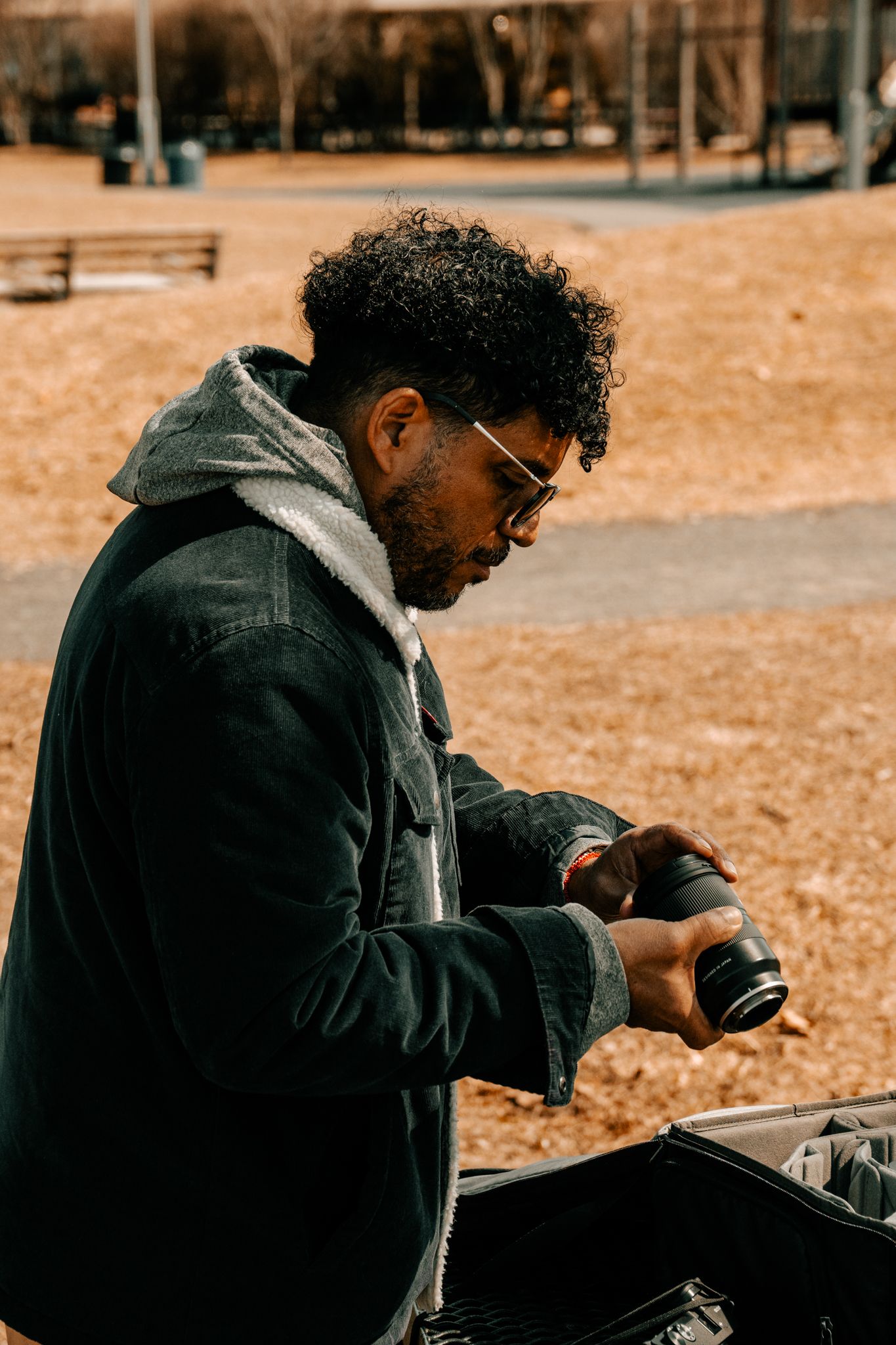 Young Black man with curly hair and glasses handling camera lens in autumn park.