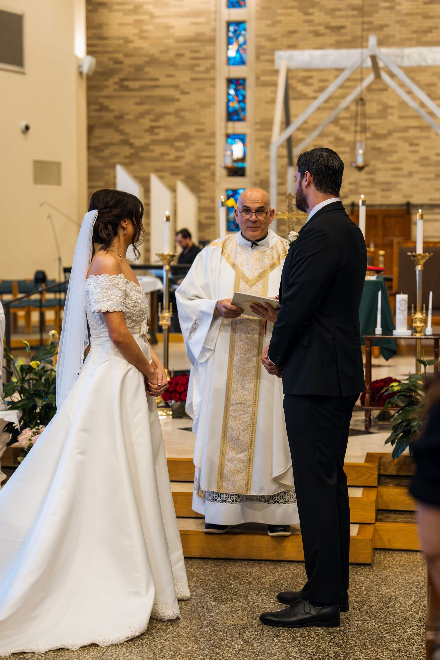 Bride and groom exchange vows before a smiling priest at a church altar during a traditional wedding ceremony.