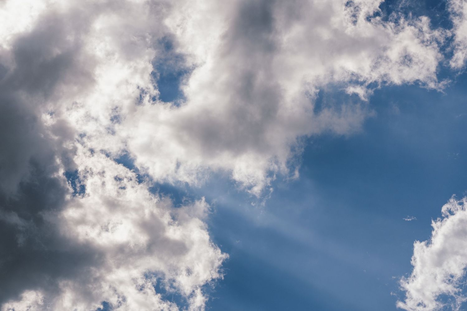 Fluffy white clouds with sun rays breaking through against a bright blue sky.