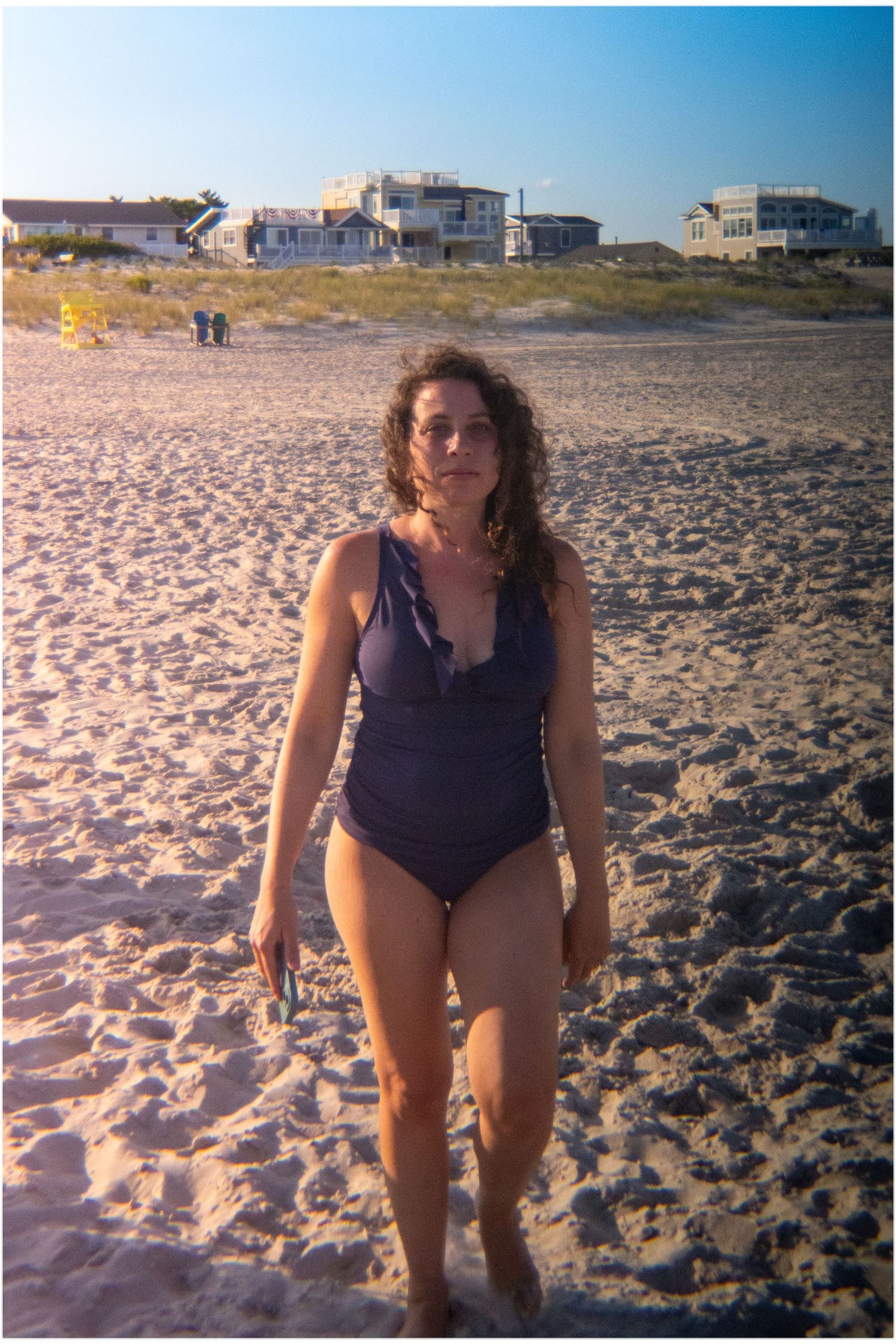 Woman in a navy one-piece swimsuit walking on a sunny beach with houses in the background.