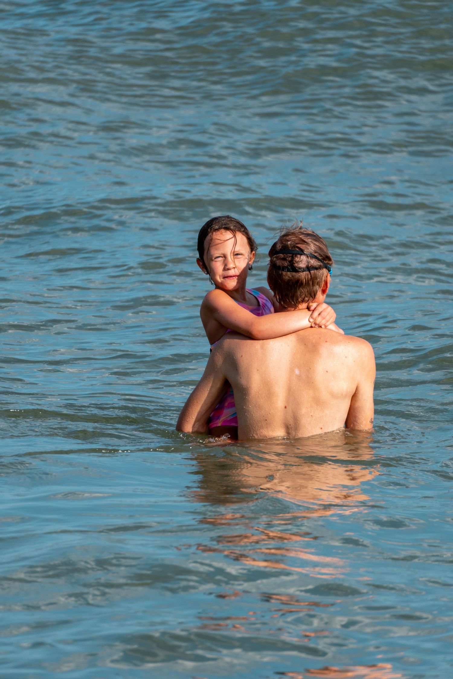 Young girl with wet hair hugging an adult from behind while wading in calm blue ocean water.