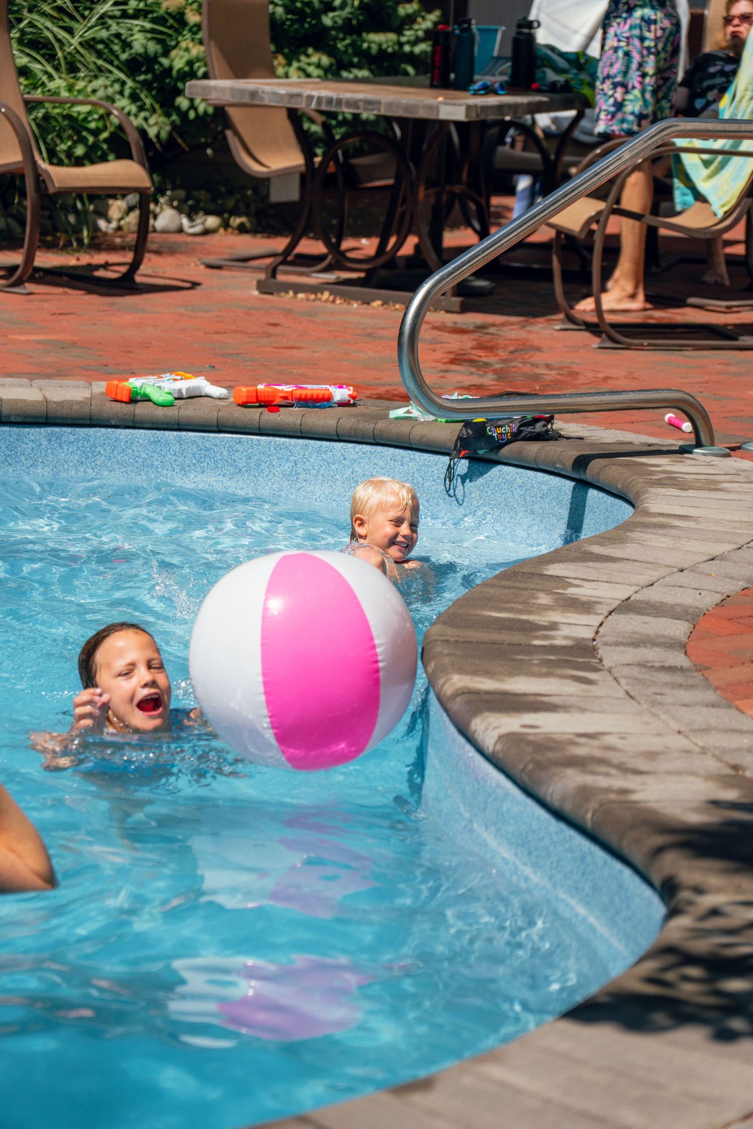Two excited children playing with a pink beach ball in a sparkling backyard swimming pool.