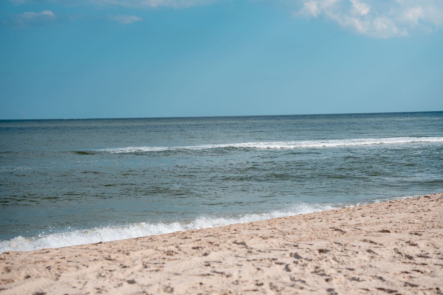 Peaceful ocean waves lapping onto a sandy shore under a bright blue partly cloudy sky.