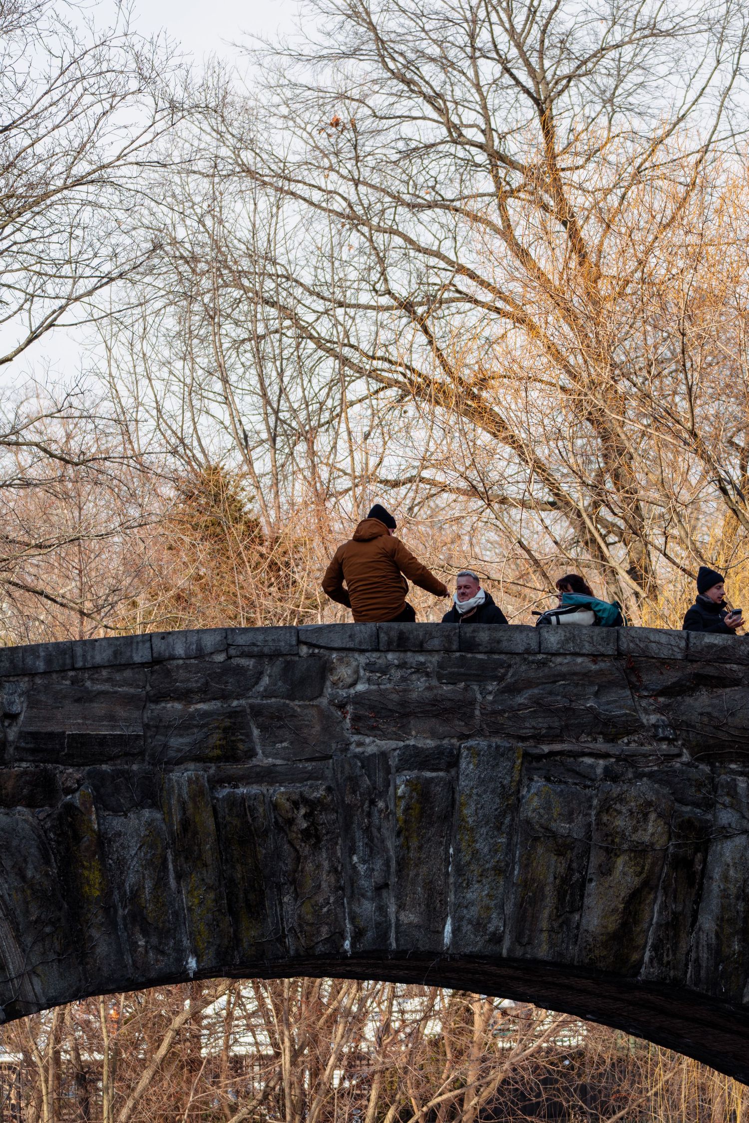 People walking on a historic stone arch bridge with bare trees in the background.