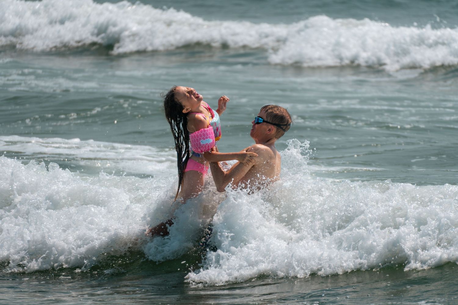 Two people playing in ocean waves at the beach, with a child being lifted by an adult amid splashing surf.