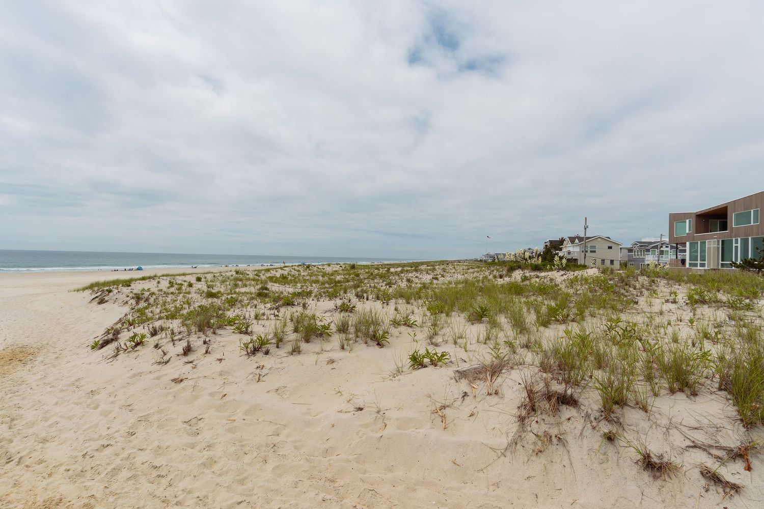 Sandy beach with coastal dunes, green vegetation, and beachfront homes under a cloudy sky.