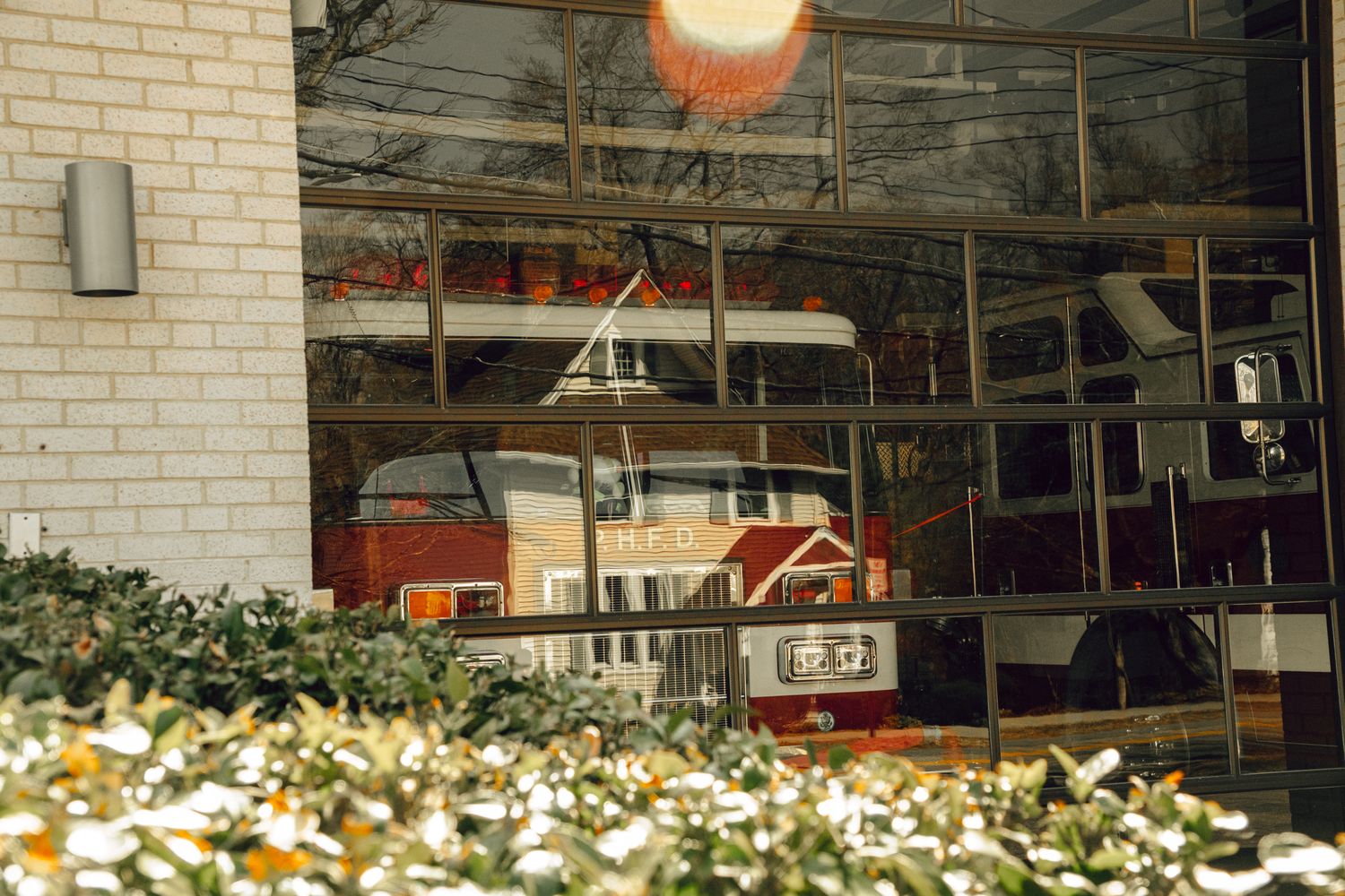 Red fire truck partially visible through reflective glass firehouse doors with white brick exterior and shrubs.