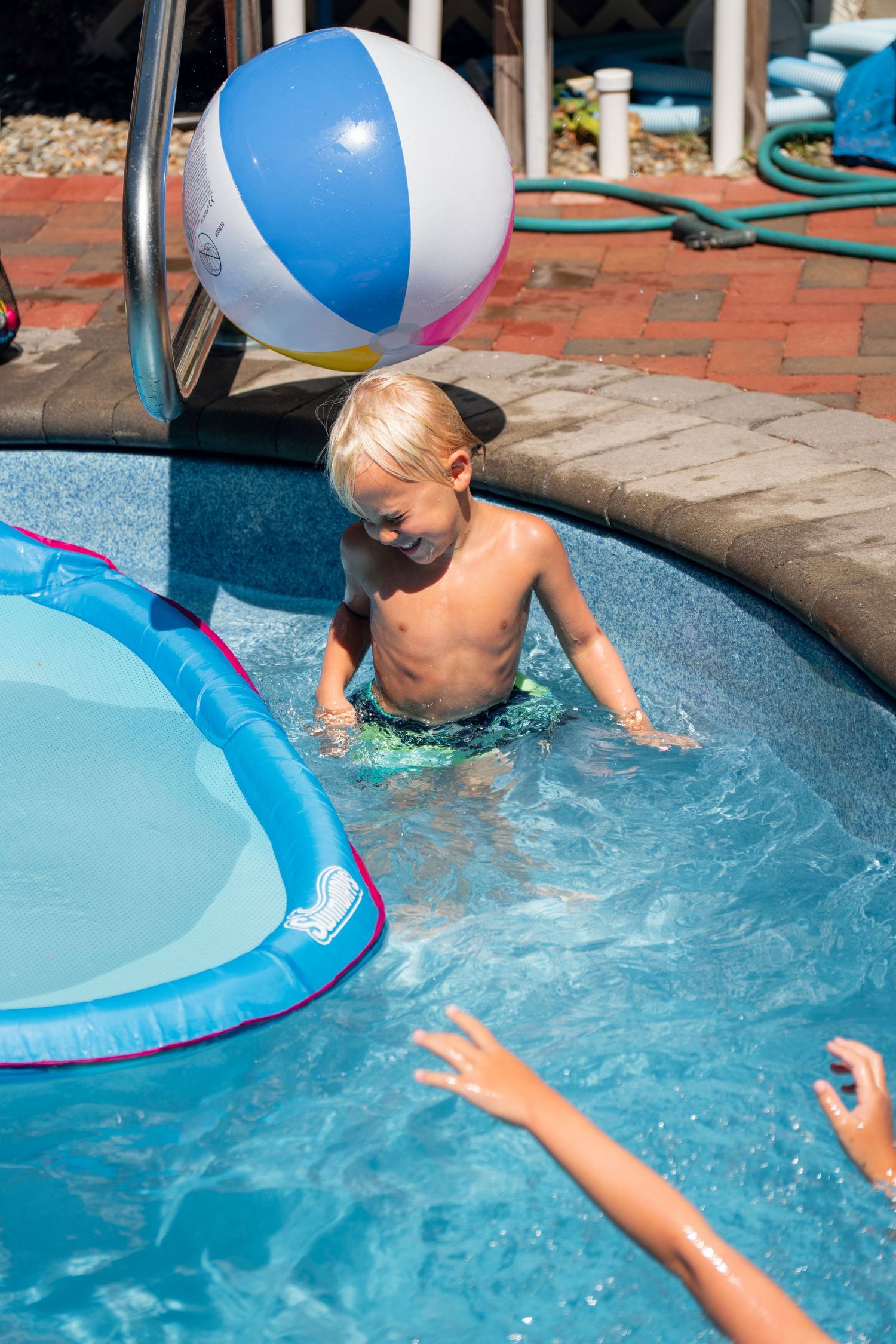 Blonde toddler standing in shallow end of backyard pool with colorful beach ball overhead.