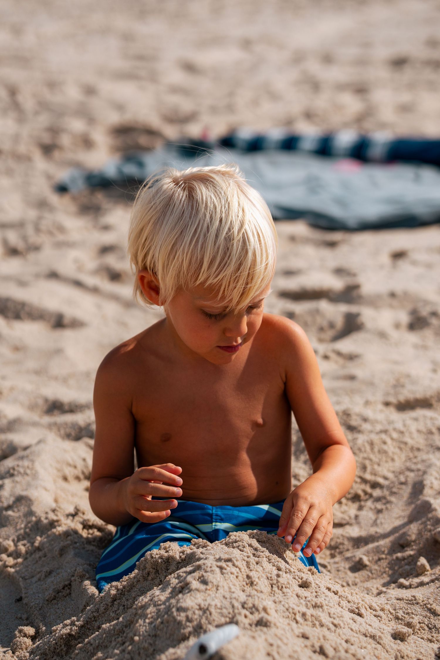Young blonde boy in blue swim trunks sitting and playing in sand at the beach.