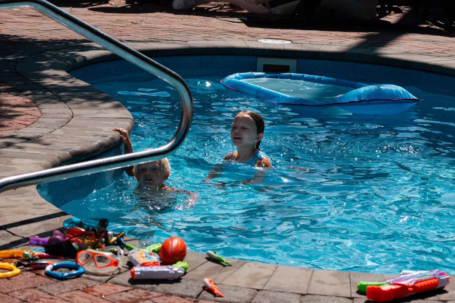 Two children swimming in a backyard pool surrounded by colorful pool toys.