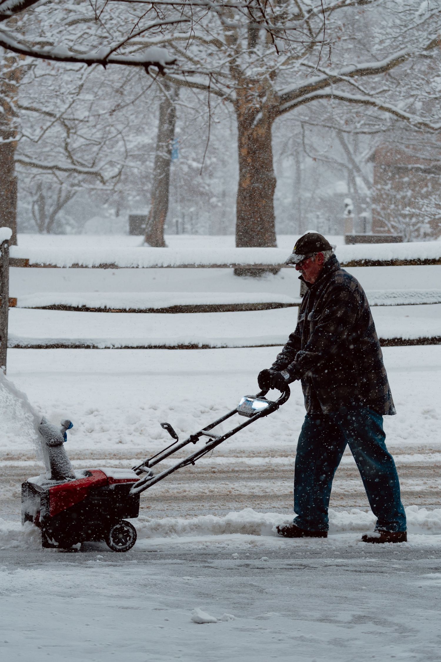 An older man pushing a red snowblower along a snowy driveway during a heavy winter snowfall.