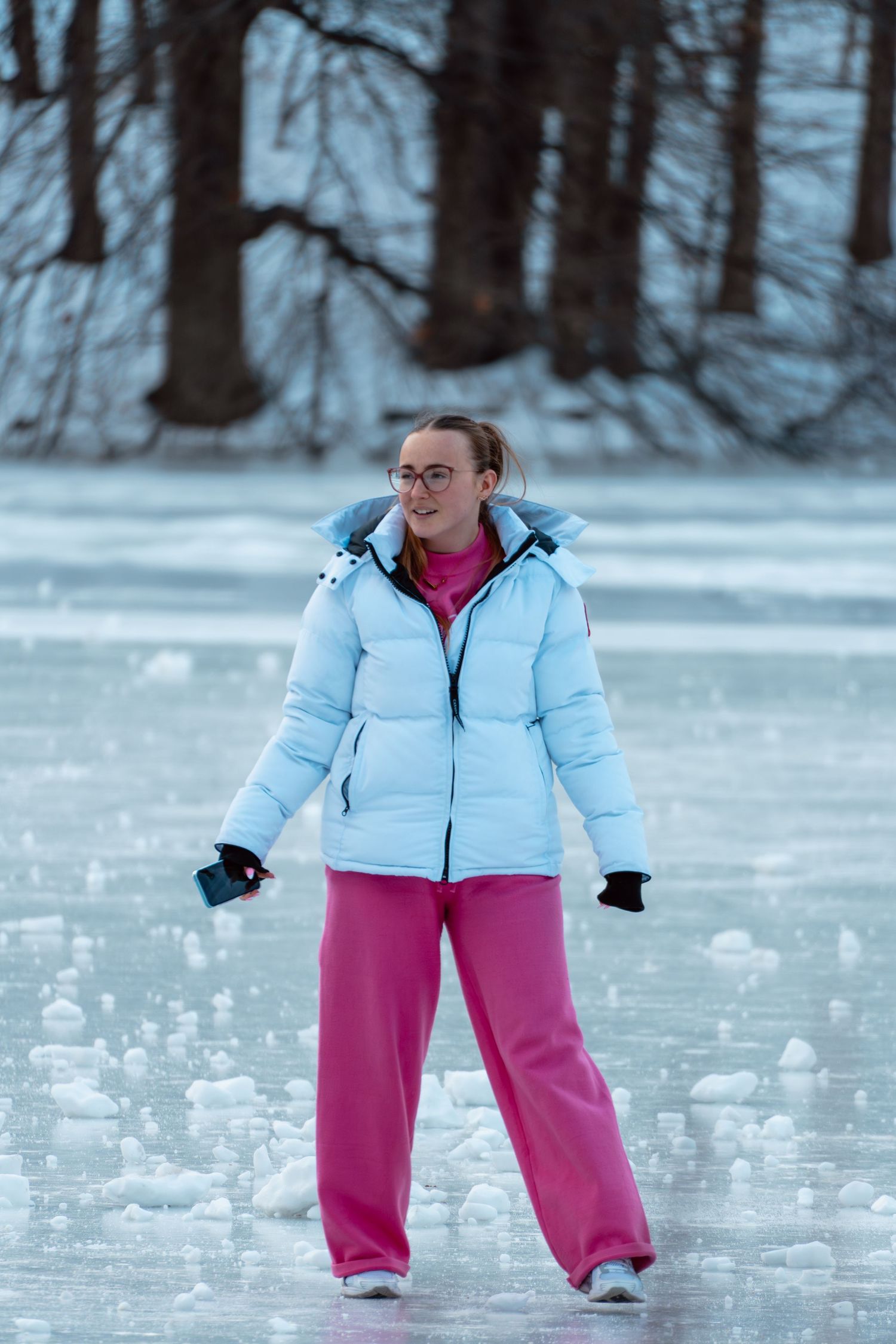 A smiling young woman wearing glasses and a light blue puffer jacket ice skates on a frozen lake.