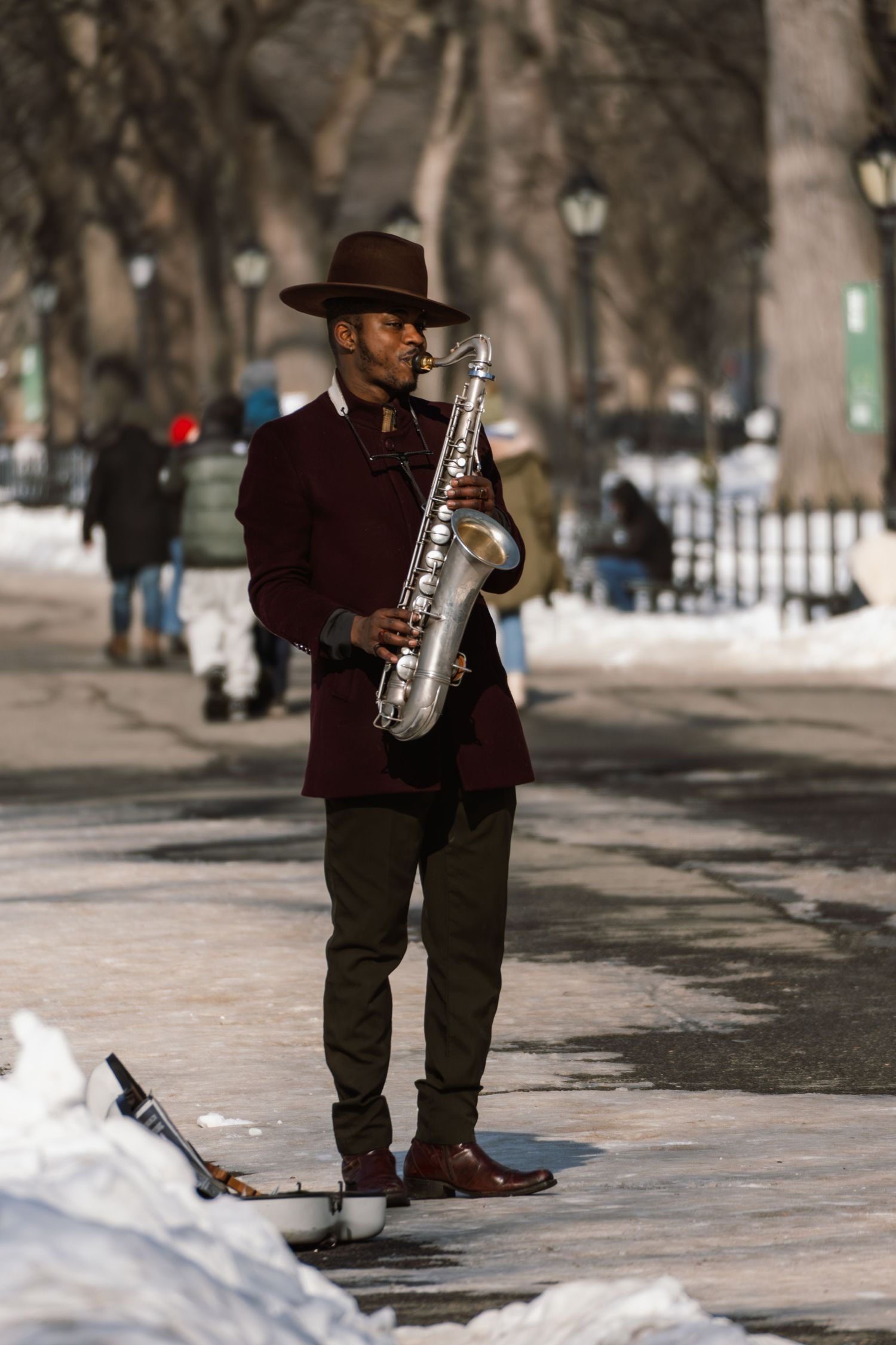 A street musician in a wide-brim hat plays saxophone on a snow-lined Central Park walkway.
