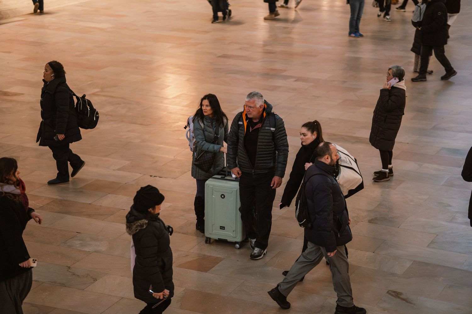 Travelers walking through Grand Central Terminal, with a couple pulling a gray suitcase through the busy concourse.