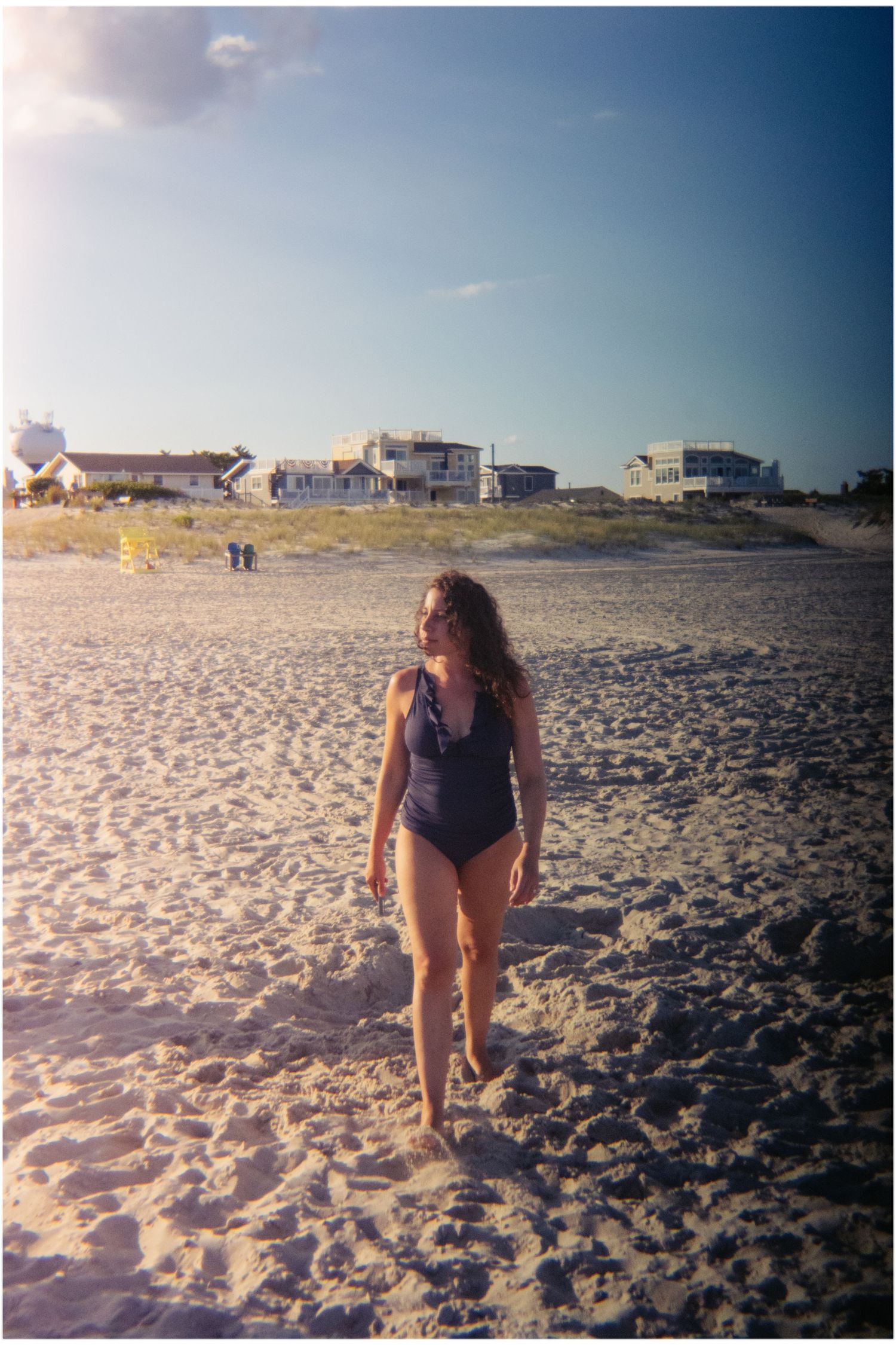 Woman in a dark swimsuit strolling on a wide sandy beach with coastal homes behind her.