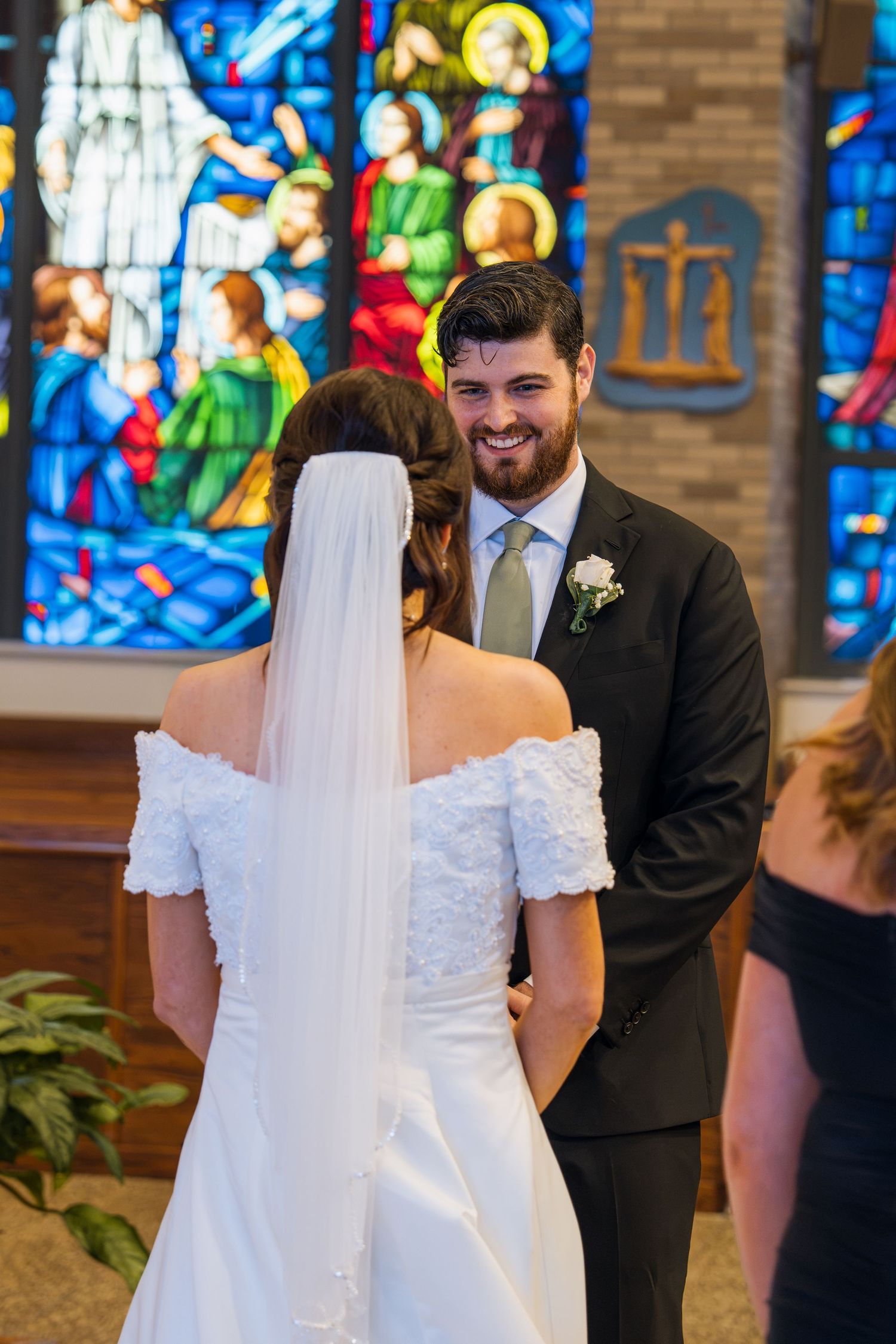 Groom smiling at bride during wedding ceremony in church with colorful stained glass windows in background.