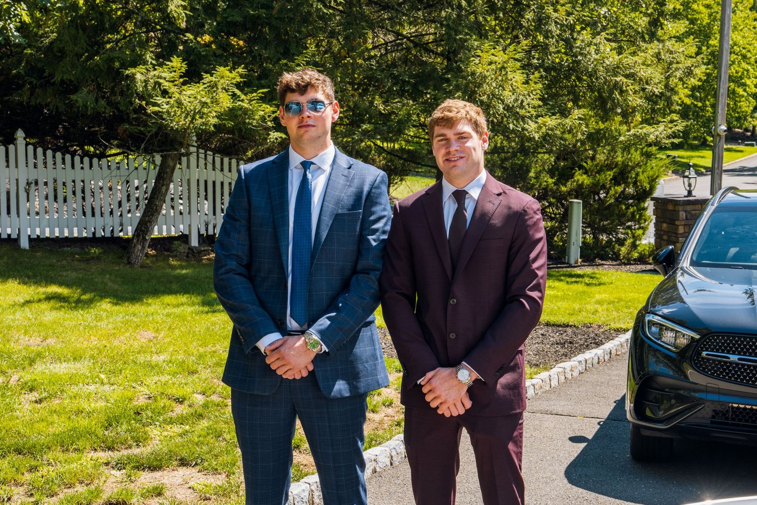 Two young men in formal suits posing outdoors on a sunny day near a white fence and luxury car.