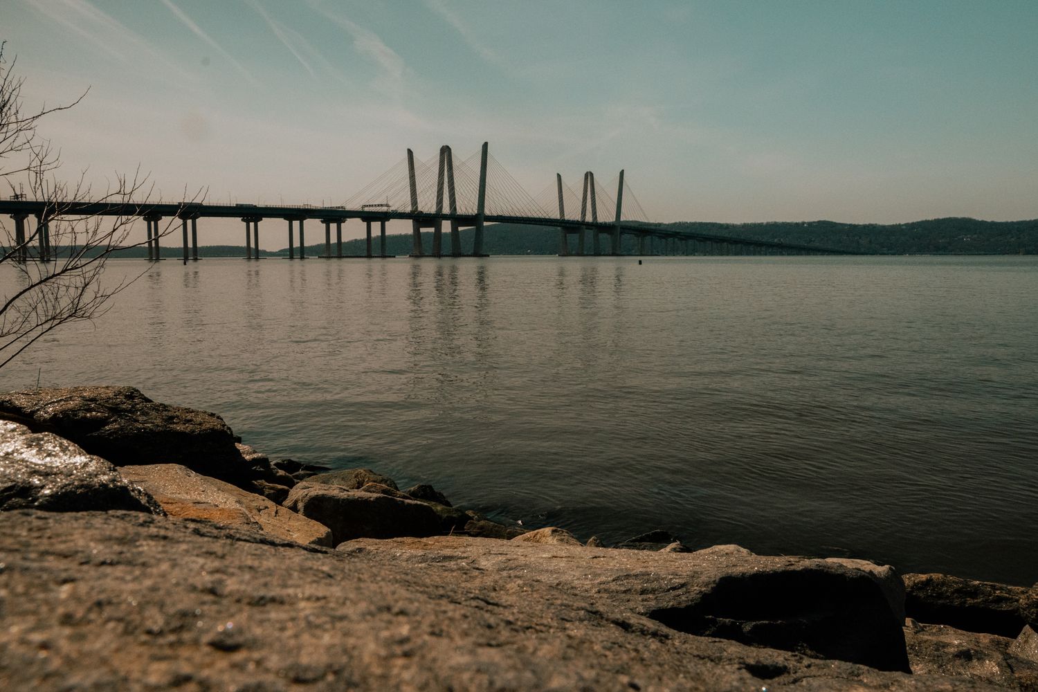 Cable-stayed bridge stretches across a wide river viewed from a rocky shore with bare winter trees nearby.