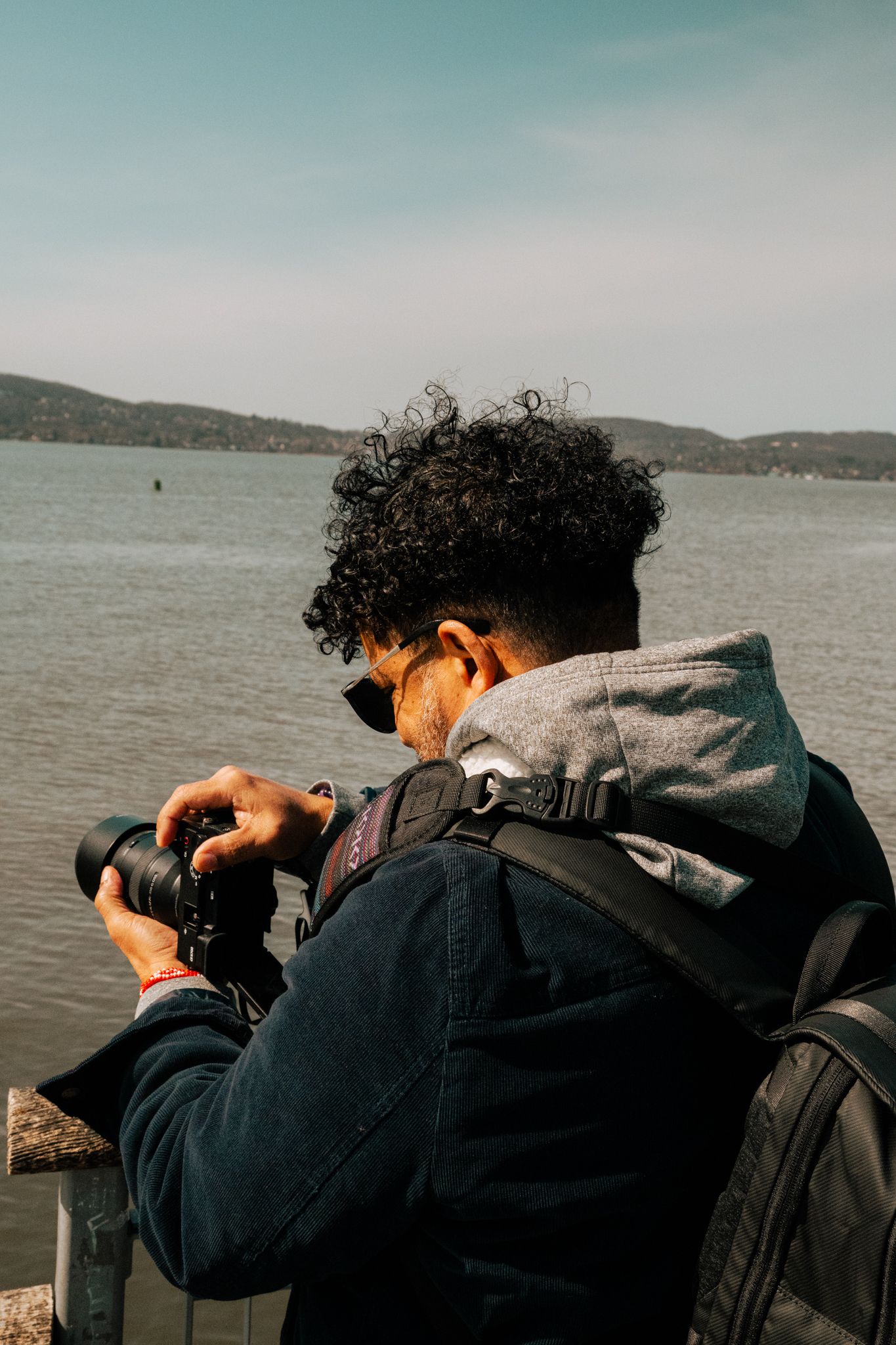 A curly-haired photographer with a backpack reviews shots on a camera beside a scenic river.