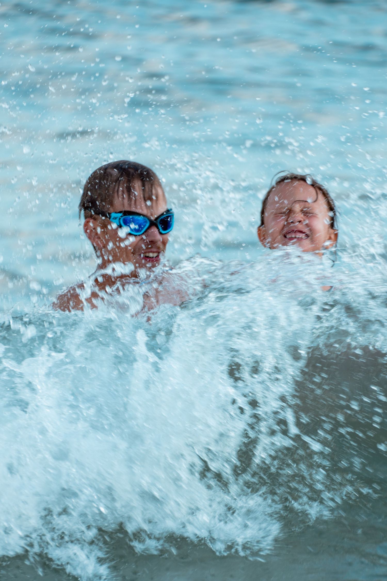 Two children caught in a splashing ocean wave, one wearing blue swimming goggles at the beach.