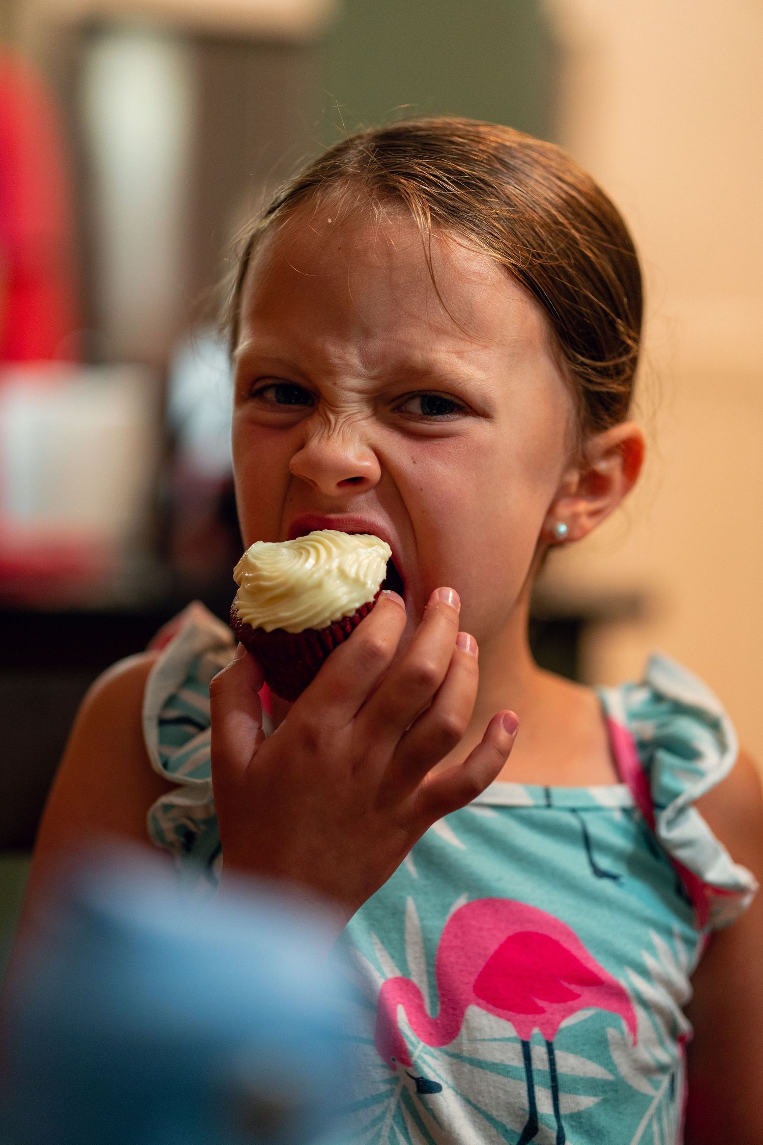 A young girl in a teal flamingo dress takes a big bite of a chocolate cupcake with white frosting.