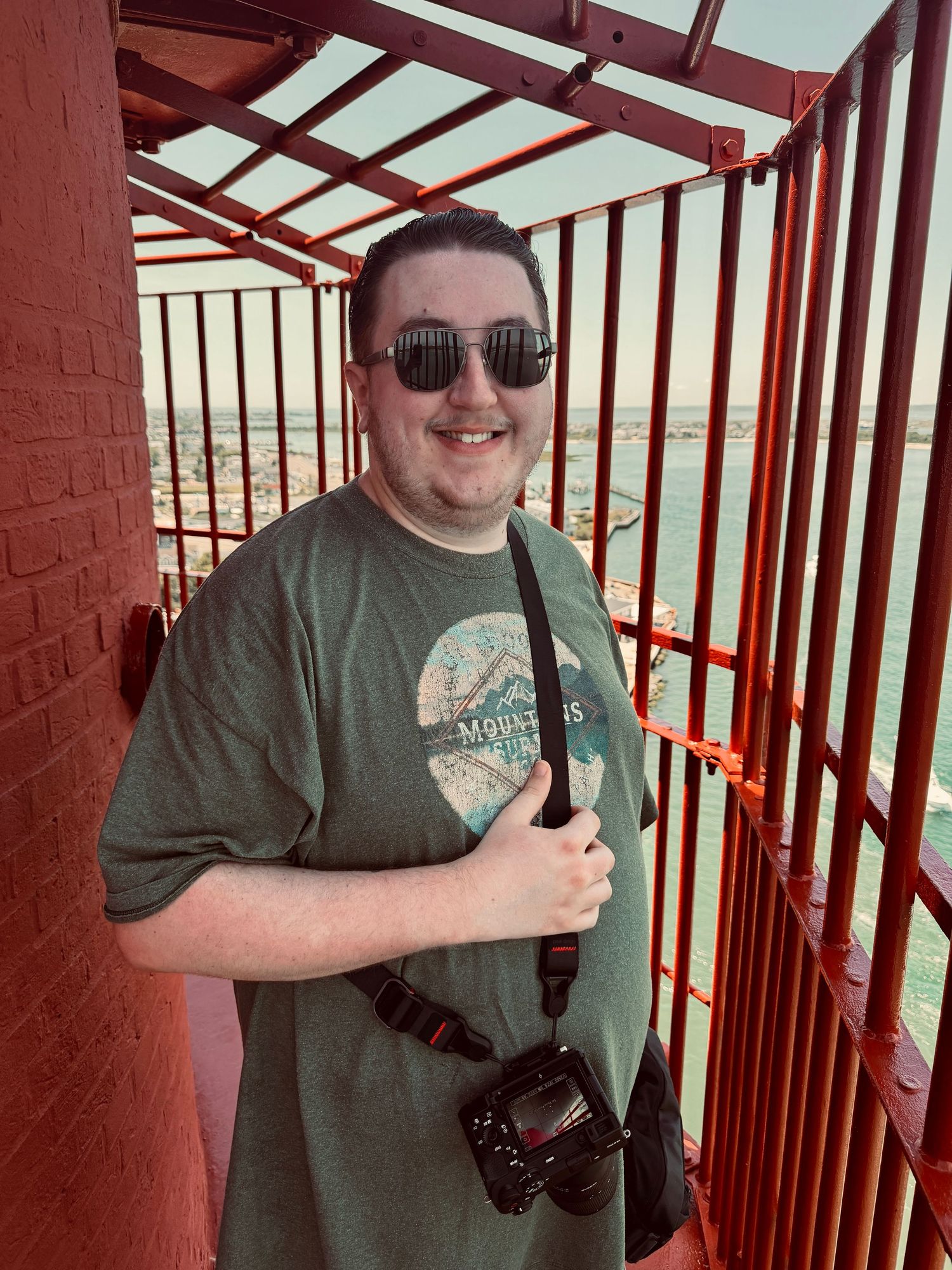 Man in green t-shirt and sunglasses holding camera, smiling on red metal observation deck with scenic view.
