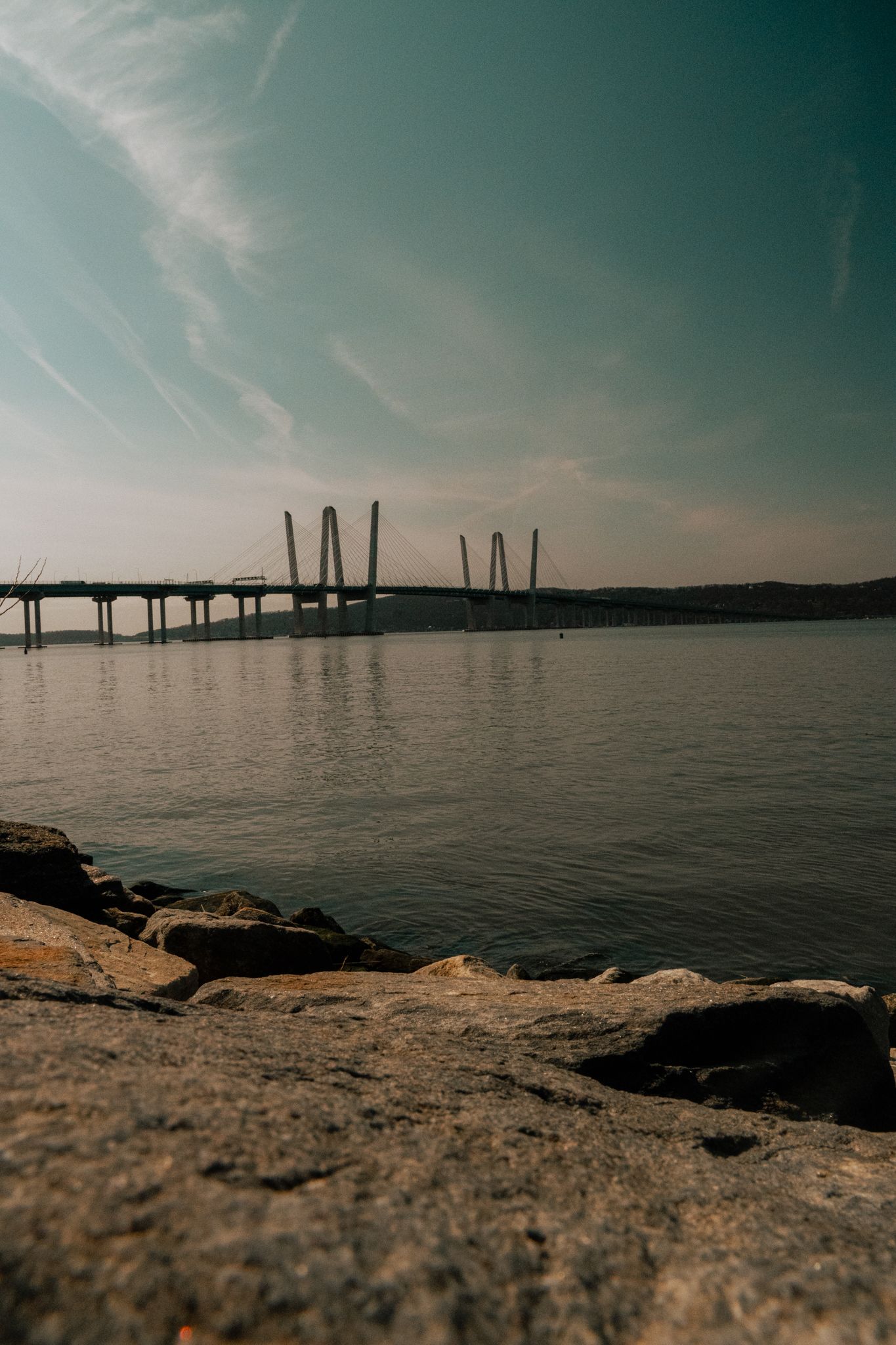Cable-stayed bridge viewed from rocky shoreline across calm reflective river water under a blue sky.