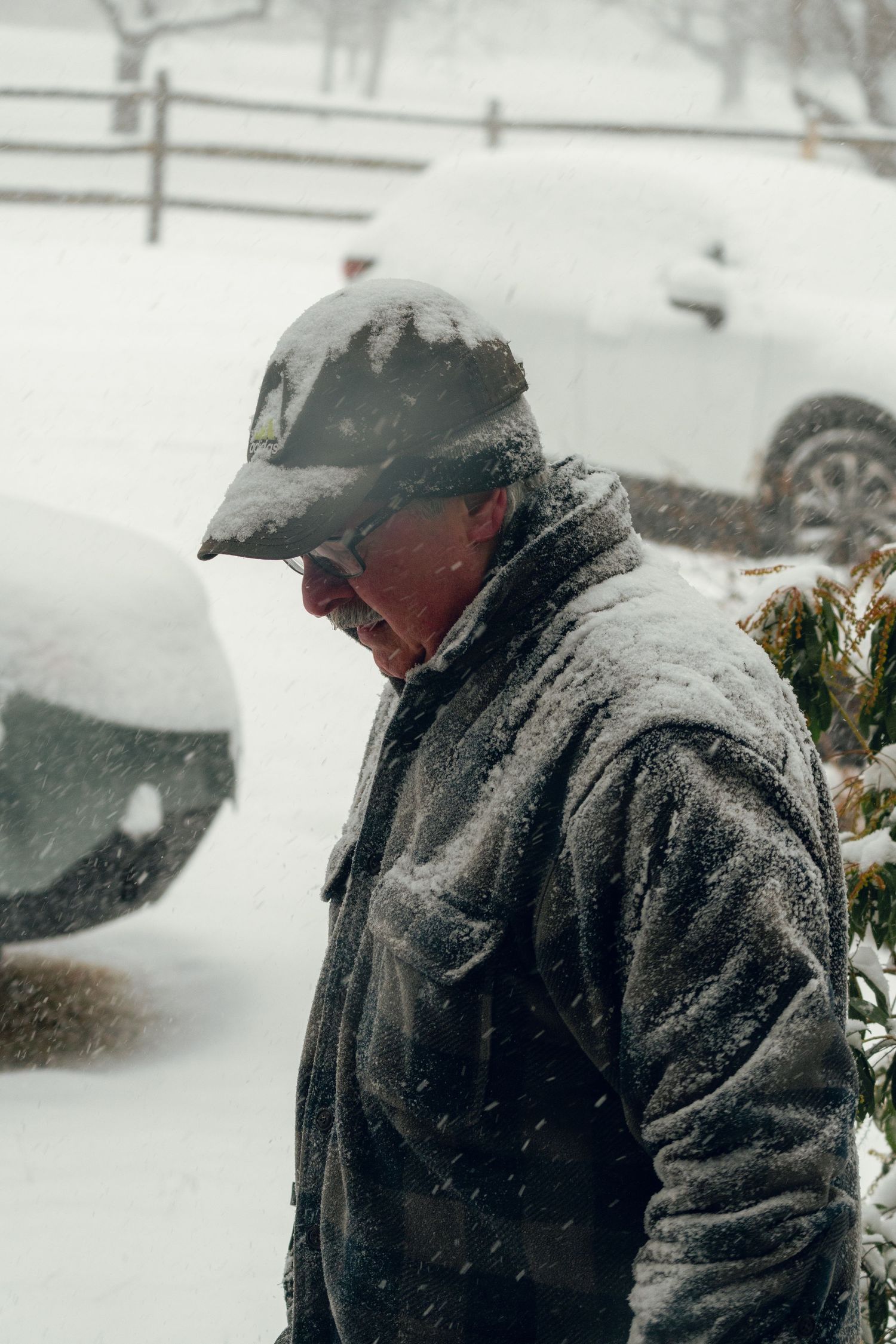 Man covered in snow during a heavy blizzard, wearing a cap and winter jacket outdoors.