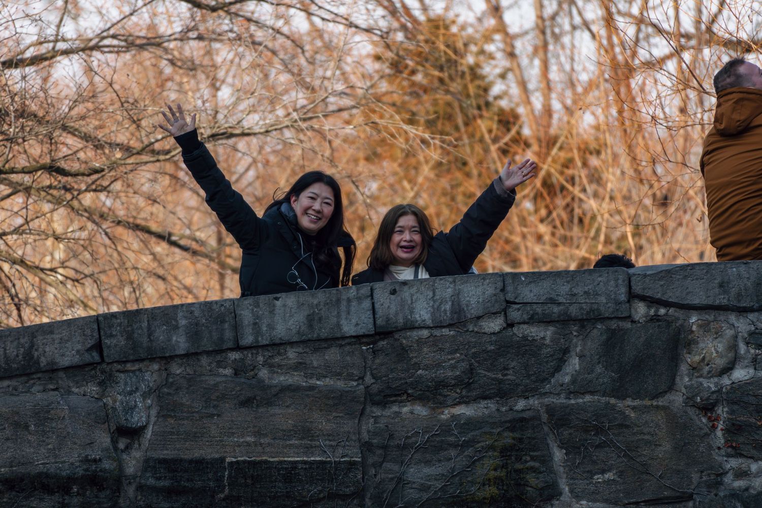 Two smiling women with arms raised joyfully pose behind a stone wall in a winter park setting.