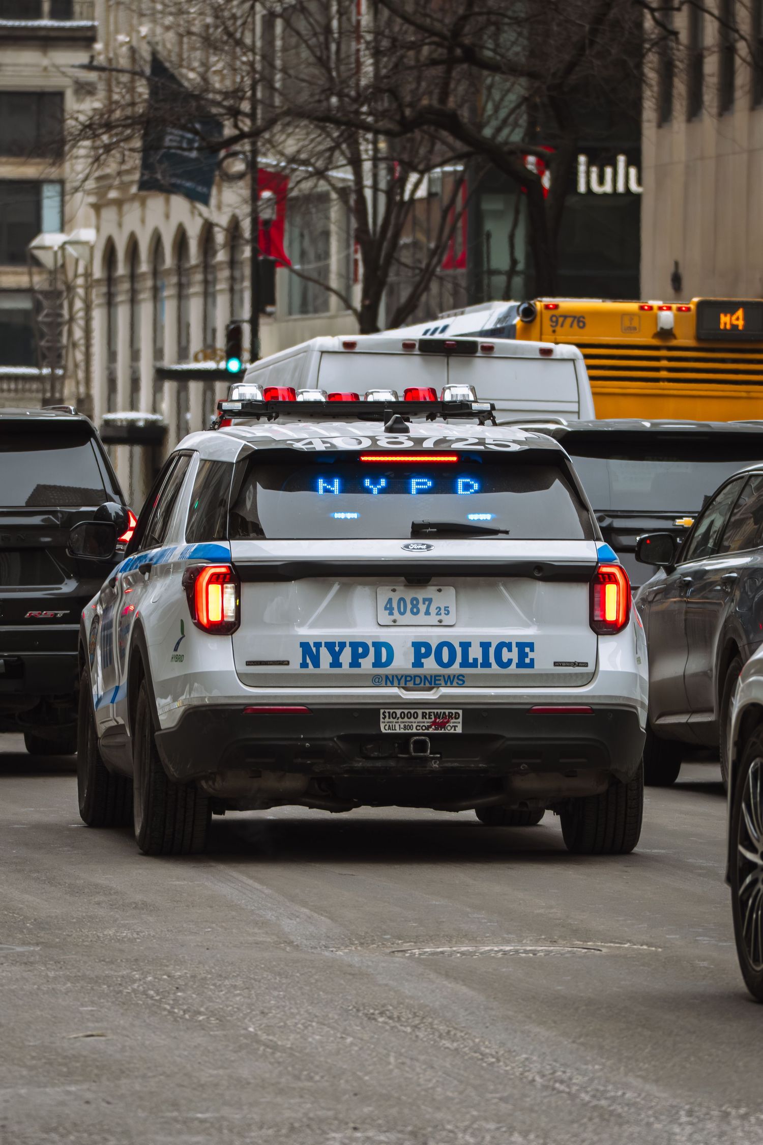 NYPD police SUV with flashing blue lights driving through midtown Manhattan traffic with a yellow school bus nearby.
