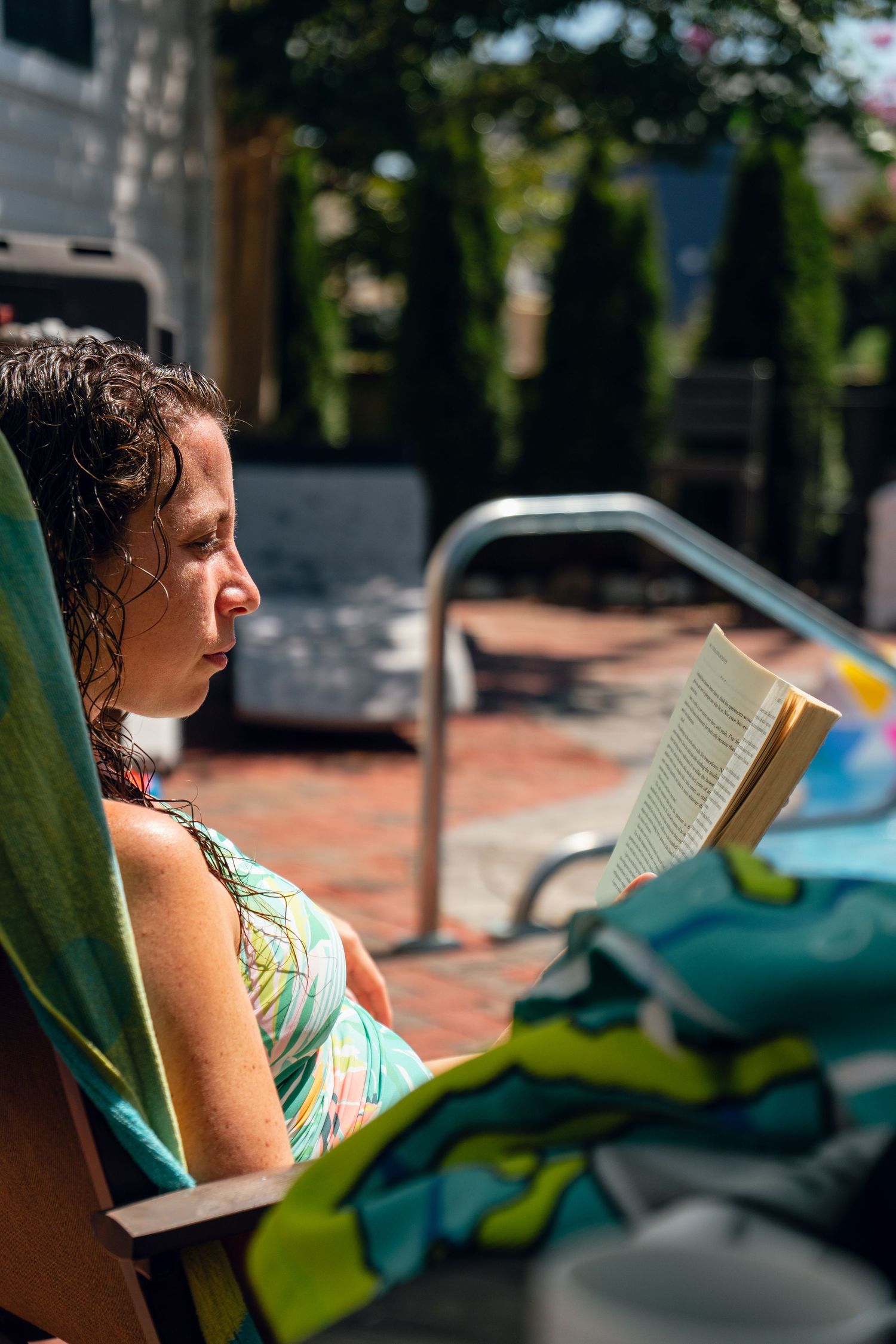 Woman in colorful swimsuit reading a book on a lounge chair beside a sunny backyard pool.