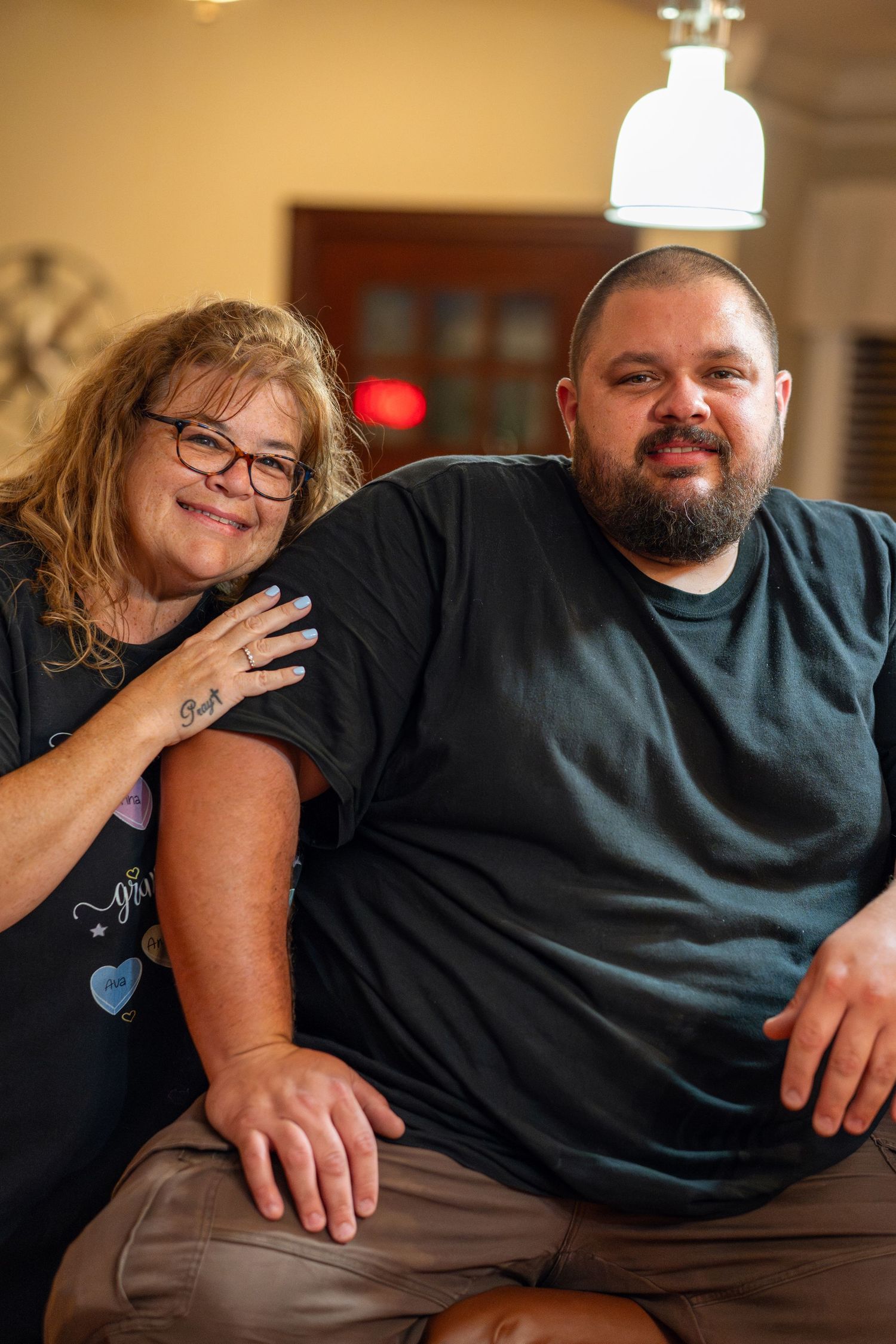 A smiling woman with glasses and a man in black t-shirt pose together warmly indoors.