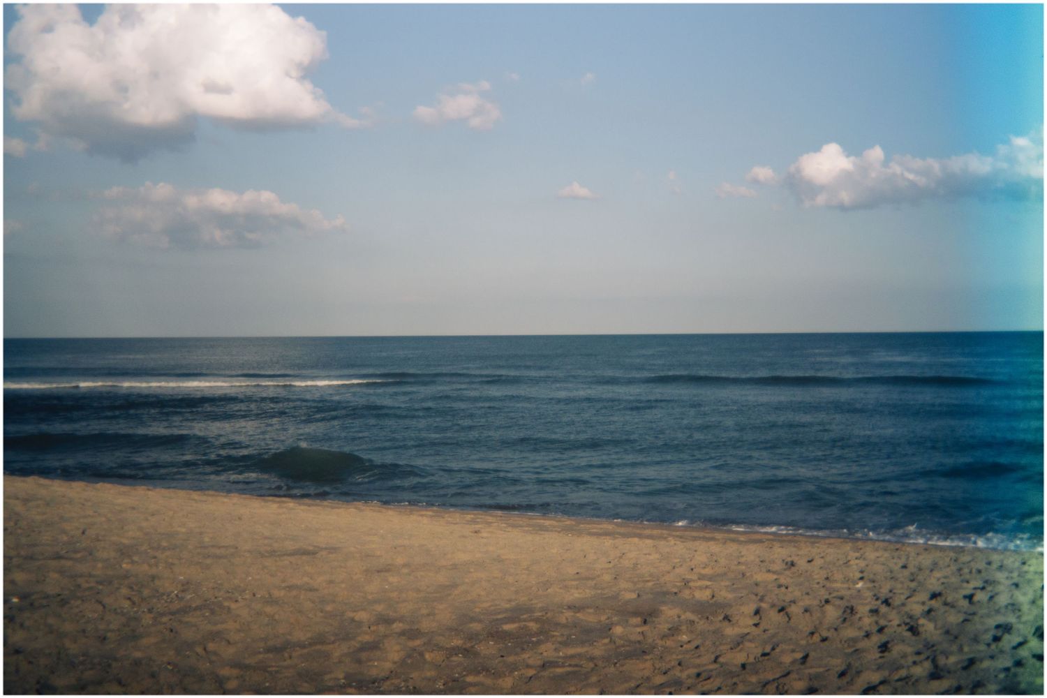 A serene empty sandy beach stretches along calm blue ocean waters under a partly cloudy sky.