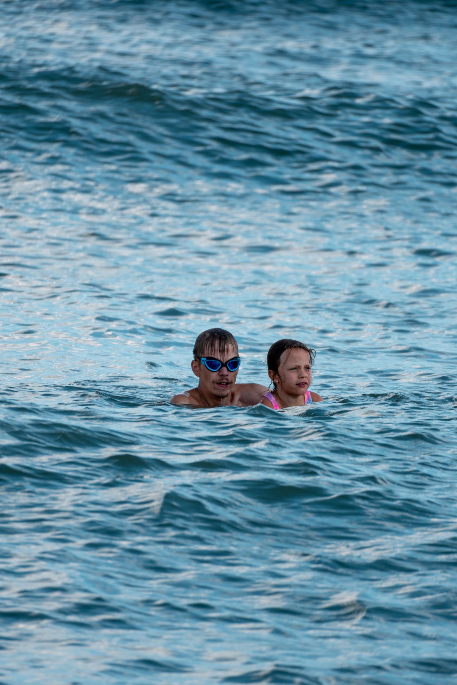 A man wearing blue goggles and a young girl swimming together in blue ocean water.