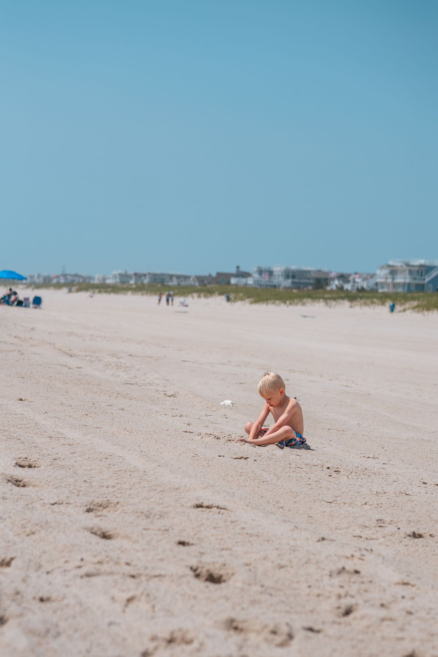 Young blonde child playing alone in white sand on a sunny beach with blue sky and distant beachfront homes.